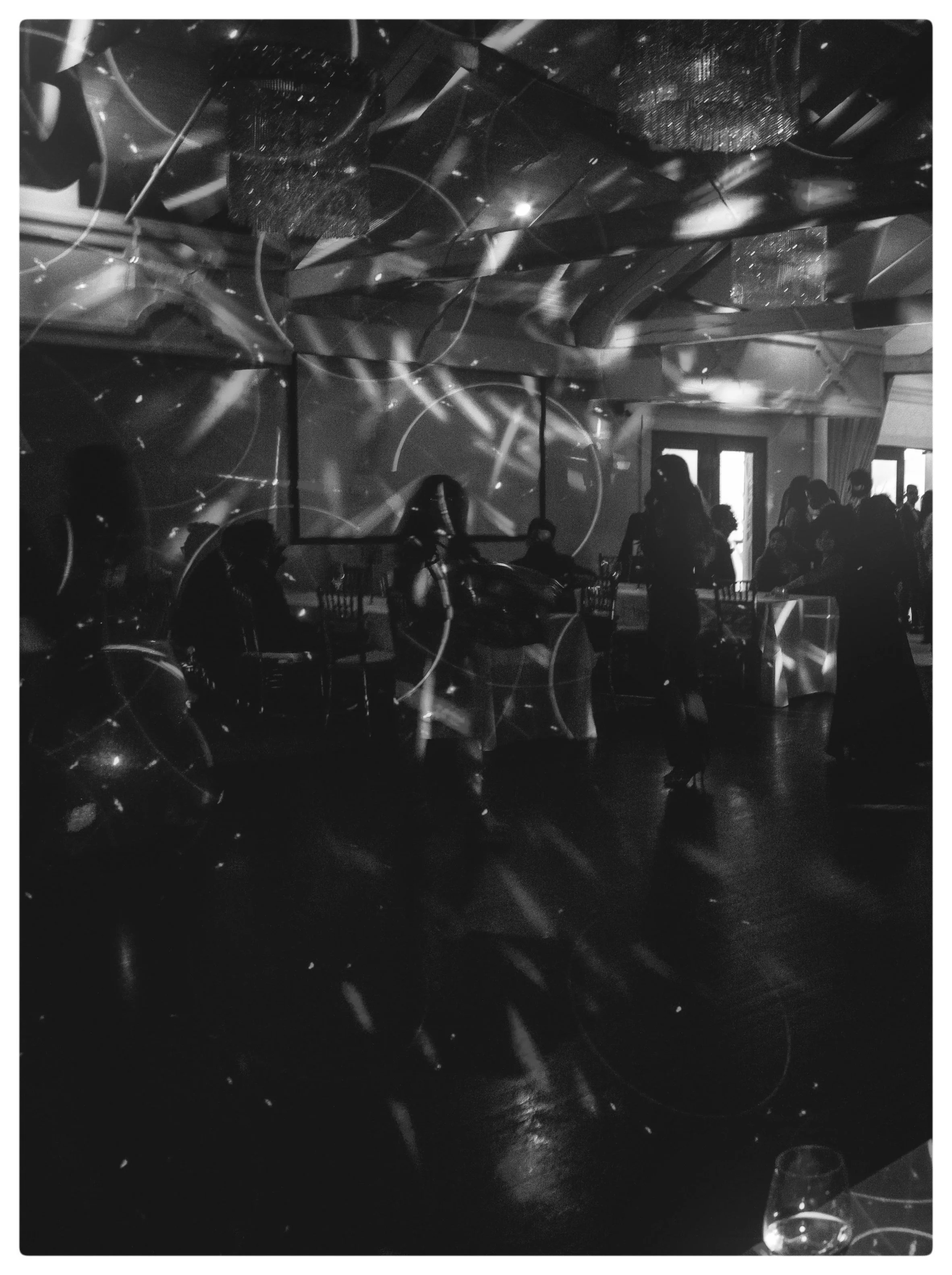 Black and white photo of people gathered in a dimly lit indoor space, possibly a restaurant or event venue, with reflections and lights on the ceiling and walls, some visible windows or doors, and tables with glasses and chairs.