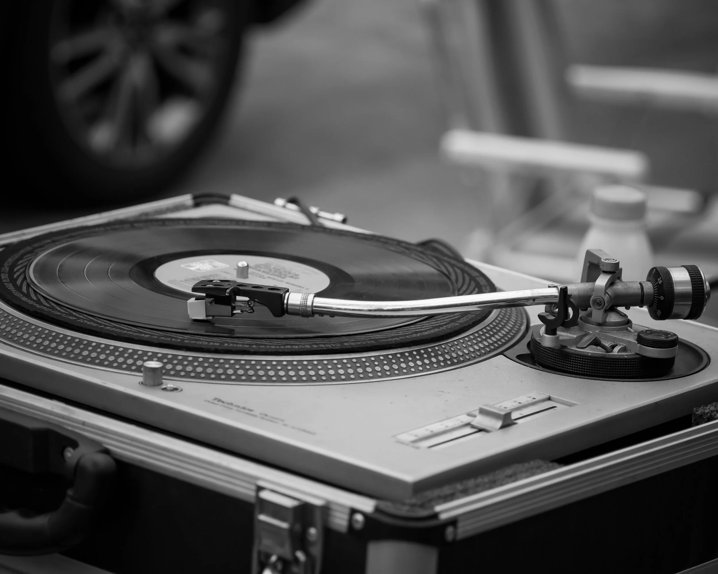 Black and white photo of a DJ turntable with a vinyl record, tonearm, and control knobs on a table, with part of a speaker visible in the background.