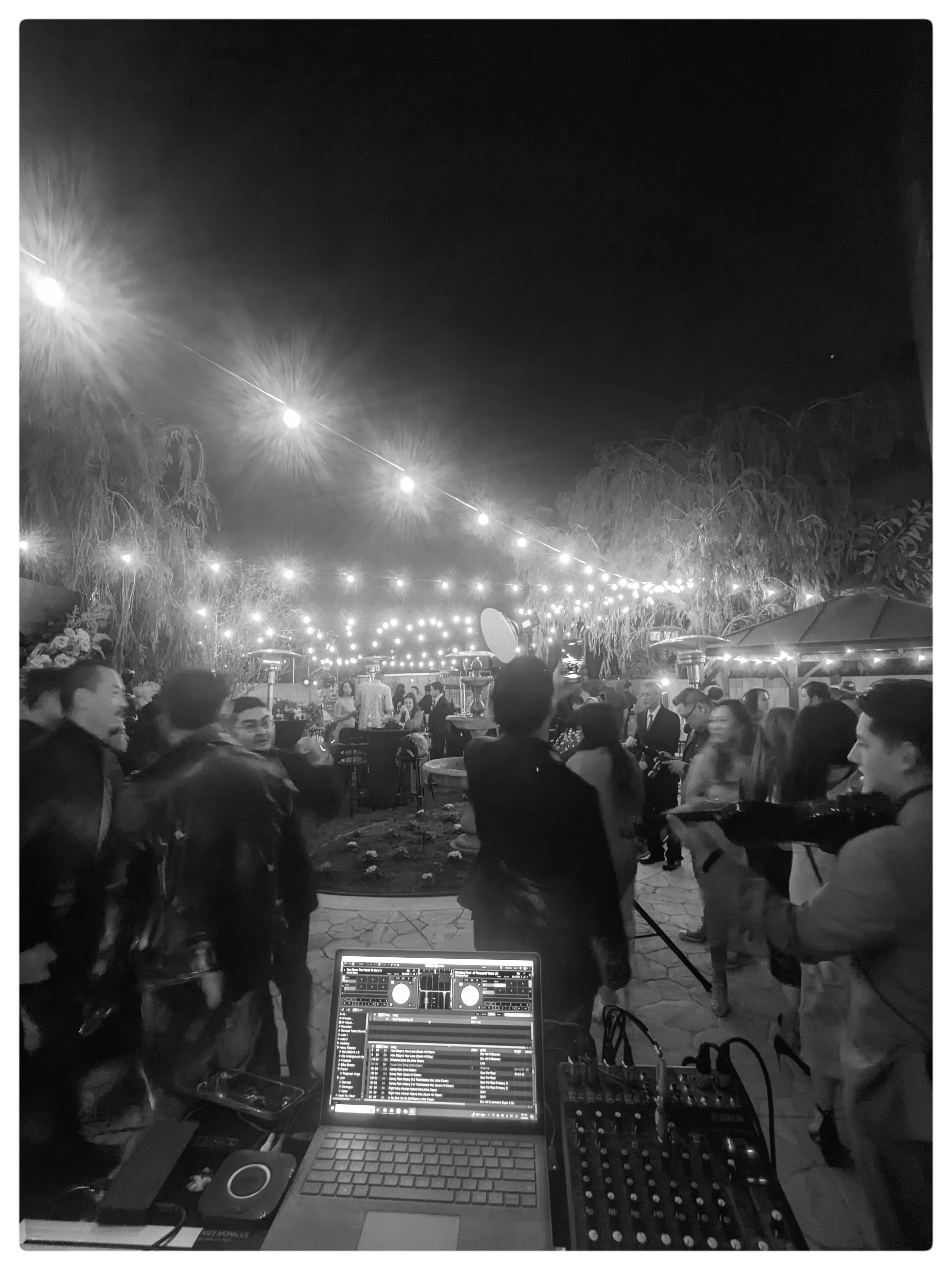 A night scene at an outdoor event with string lights hanging above and people socializing and dancing underneath. A DJ setup with a laptop and sound equipment is visible in the foreground.