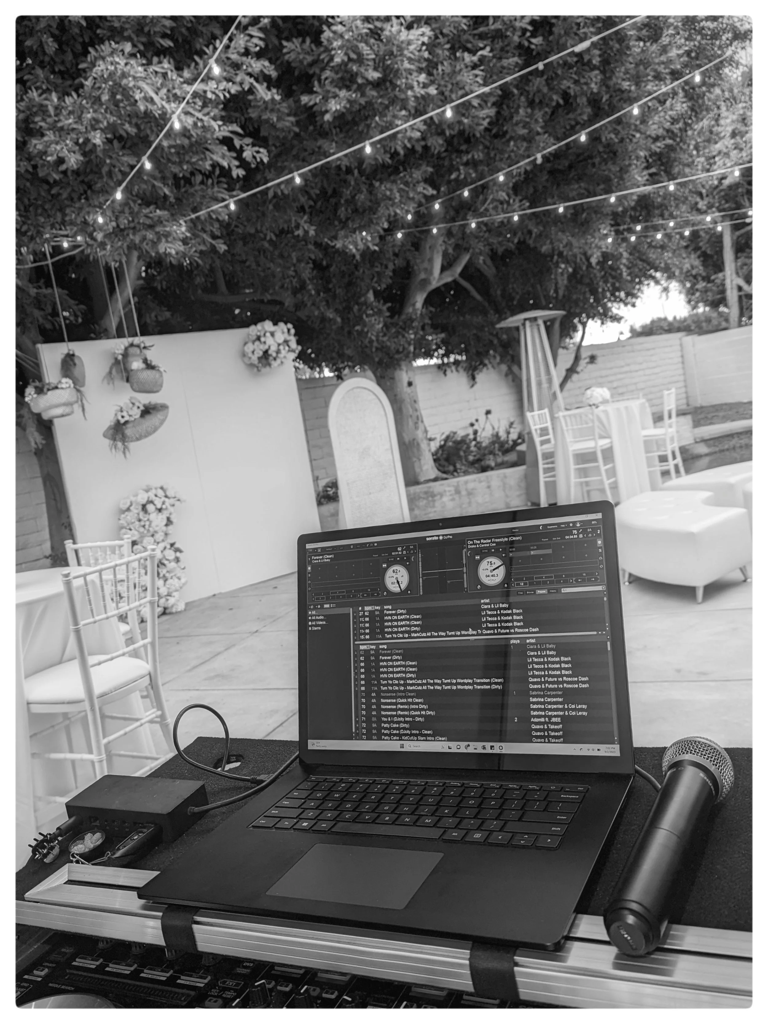A laptop on a table displaying music playlist and DJ software, with a microphone, headphones, and event decorations including string lights, flower arrangements, and hanging hats in an outdoor setting with trees and chairs.