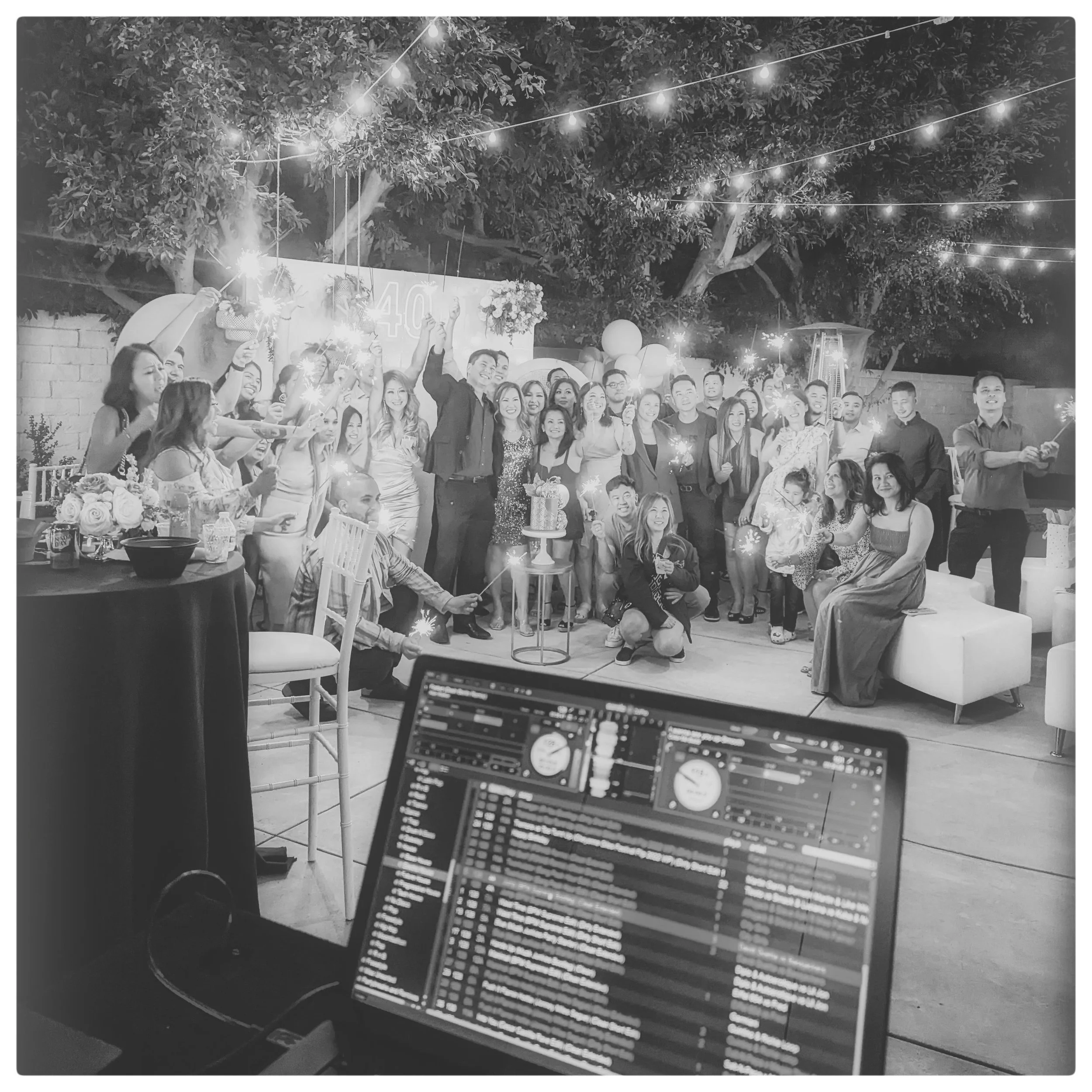 A group of people celebrating with sparklers outdoors at night under string lights, with decorations and a cake on a table, and a DJ mixing music in the foreground.