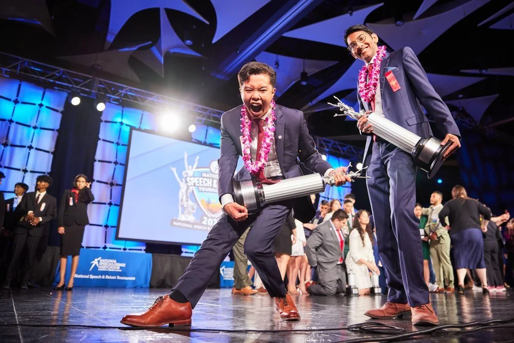 Two young men in suits holding large trophies on stage at a speech competition, celebrating with leis around their necks, with other participants and an audience in the background.