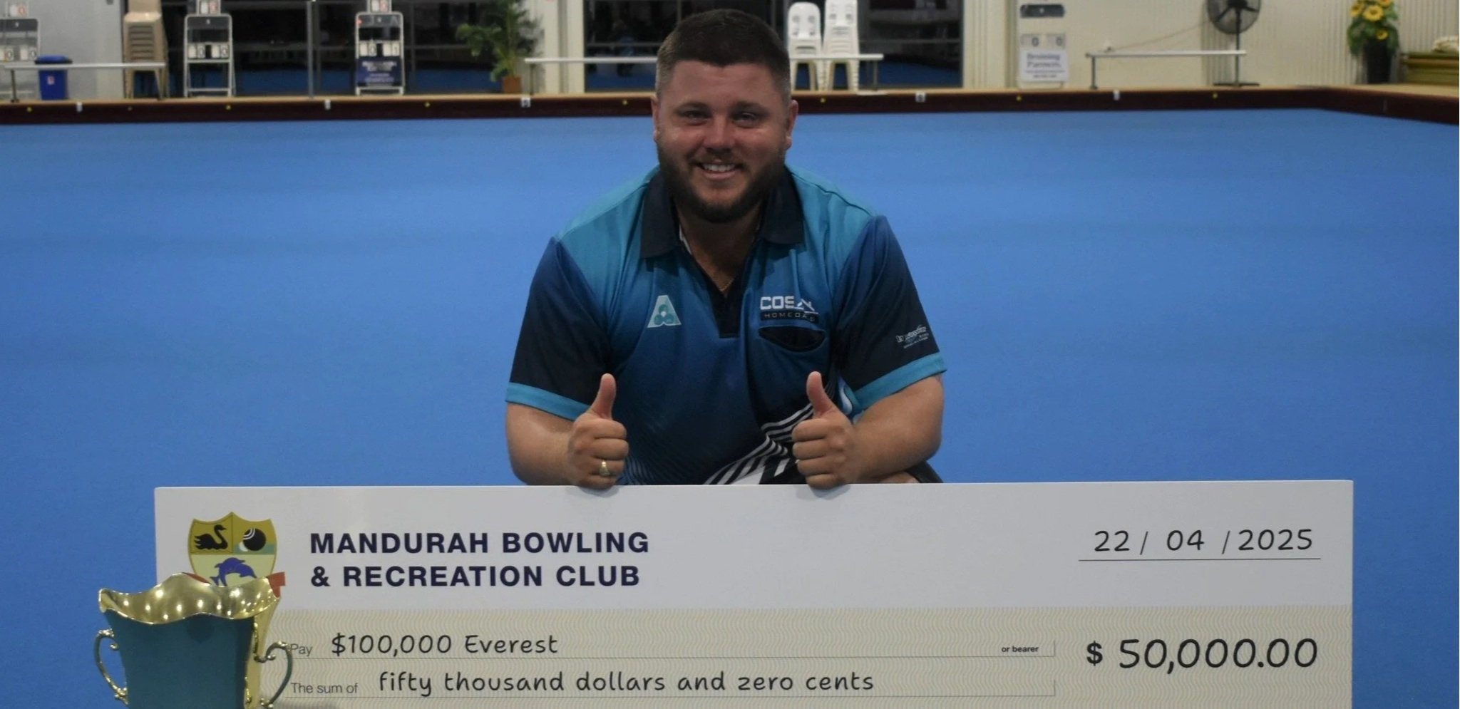 A man in a blue rowing shirt smiling and giving two thumbs up in front of a large ceremonial check from Mandurah Bowling & Recreation Club. The check is for fifty thousand dollars, dated April 22, 2025, and labeled for Everest.