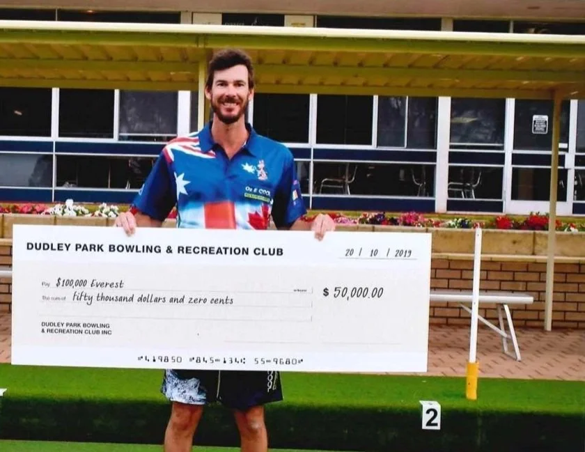 A man standing at a bowling alley holding a giant check for $50,000 from Dudley Park Bowling & Recreation Club, dated October 20, 2019, with a smile. He is wearing a colorful shirt with an American flag pattern.