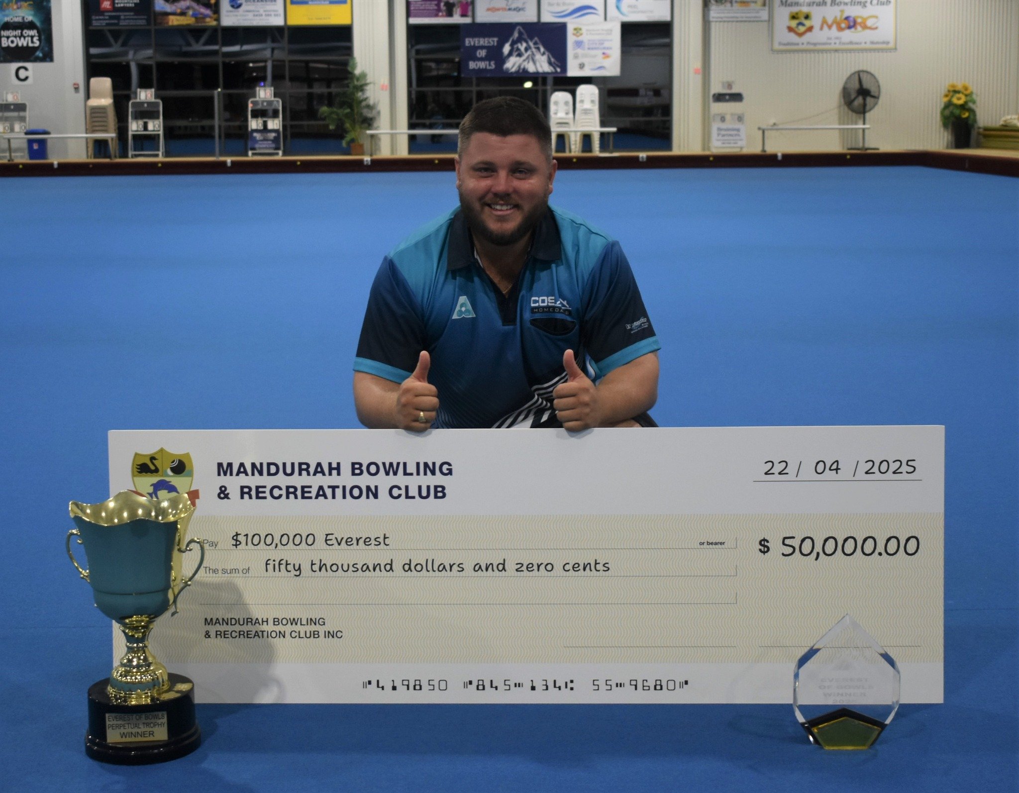 A man in a blue bowling shirt kneeling behind a large check and a trophy on a bowling alley lane. The check is from Mandurah Bowling & Recreation Club, dated 22/04/2025, payable for fifty thousand dollars, and labeled as the Everest of Bowls Perpetual Trophy Winner.