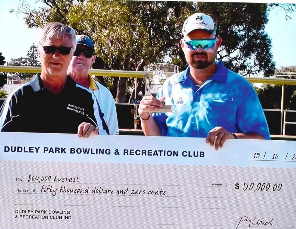 Three men at Dudley Park Bowling and Recreation Club, one holding a check for $50,000, with trees and a sports field in the background.