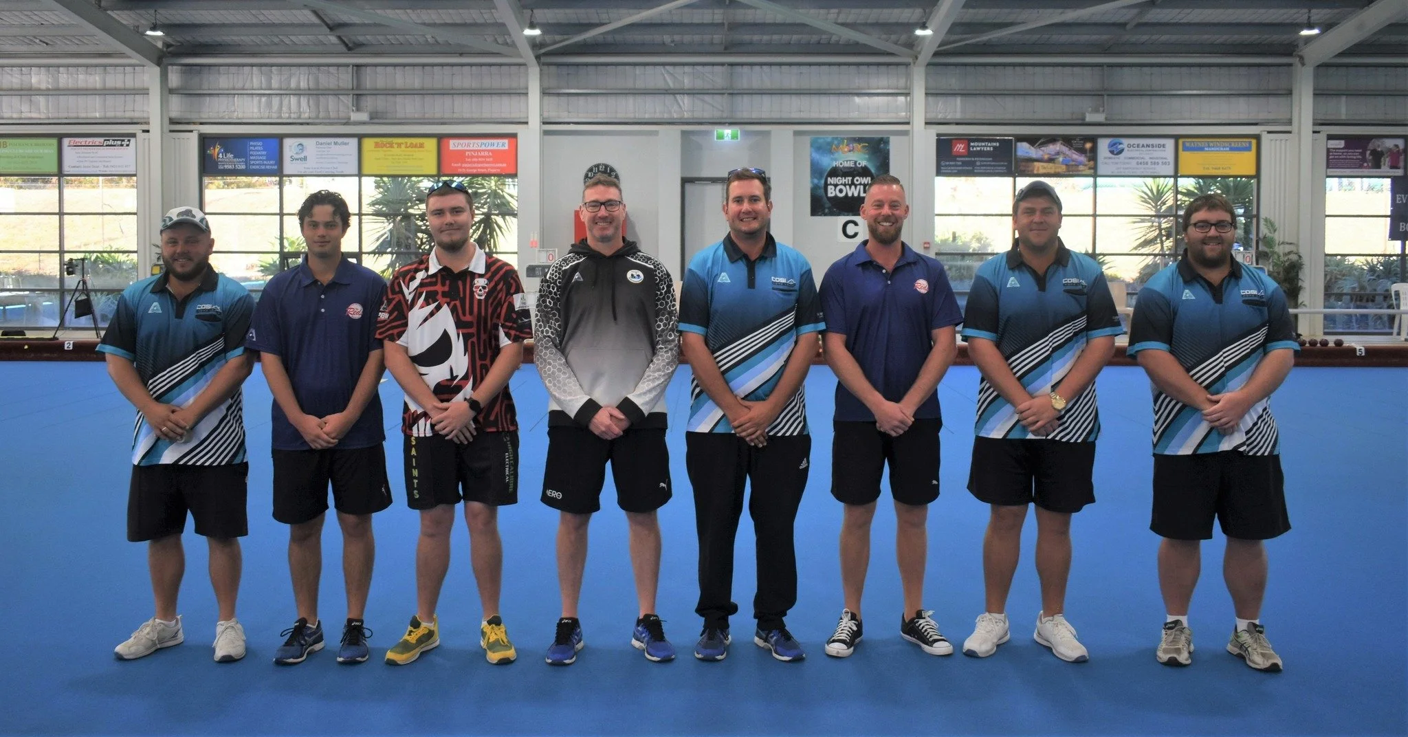 Group of eight men standing together on an indoor sports court, posing for a photo. They are wearing sports uniforms and casual clothing. The background shows large windows and advertisements.