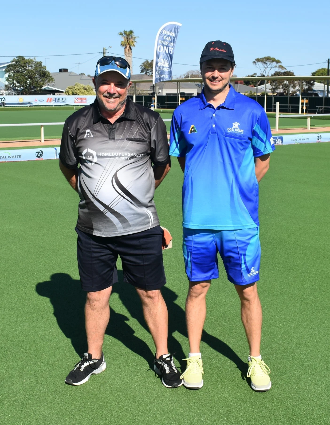 Two men standing on a bowling green, smiling, with a flag and trees in the background.