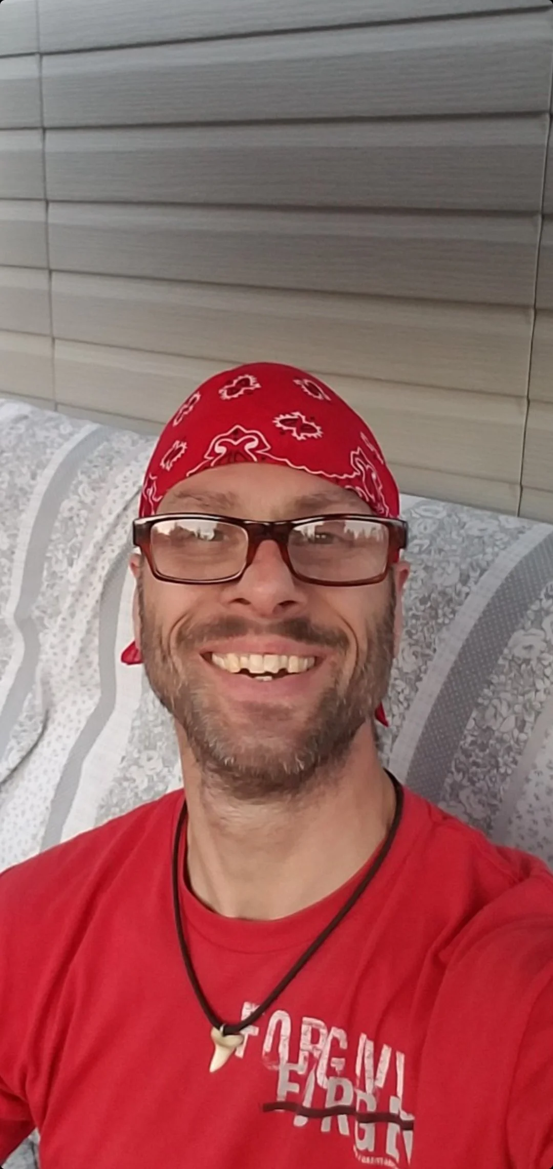 A man wearing glasses, a red bandana, and a red T-shirt, smiling while lying on a porch swing.