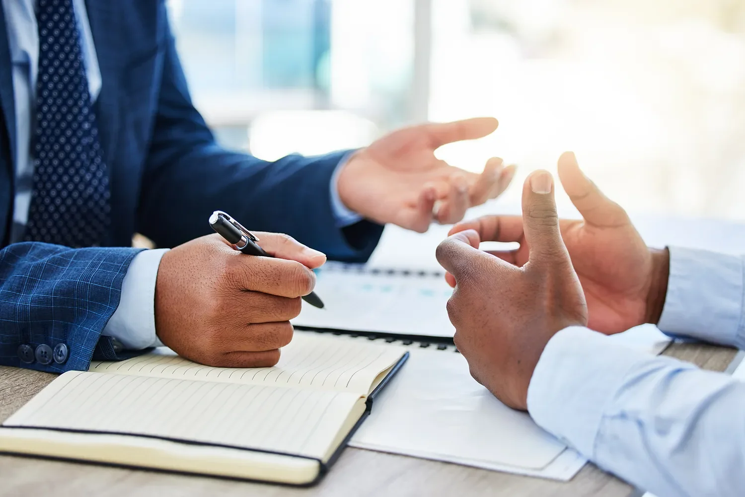 Two business professionals in suits engaged in a discussion at a table, with one taking notes with a pen and the other gesturing with their hands.