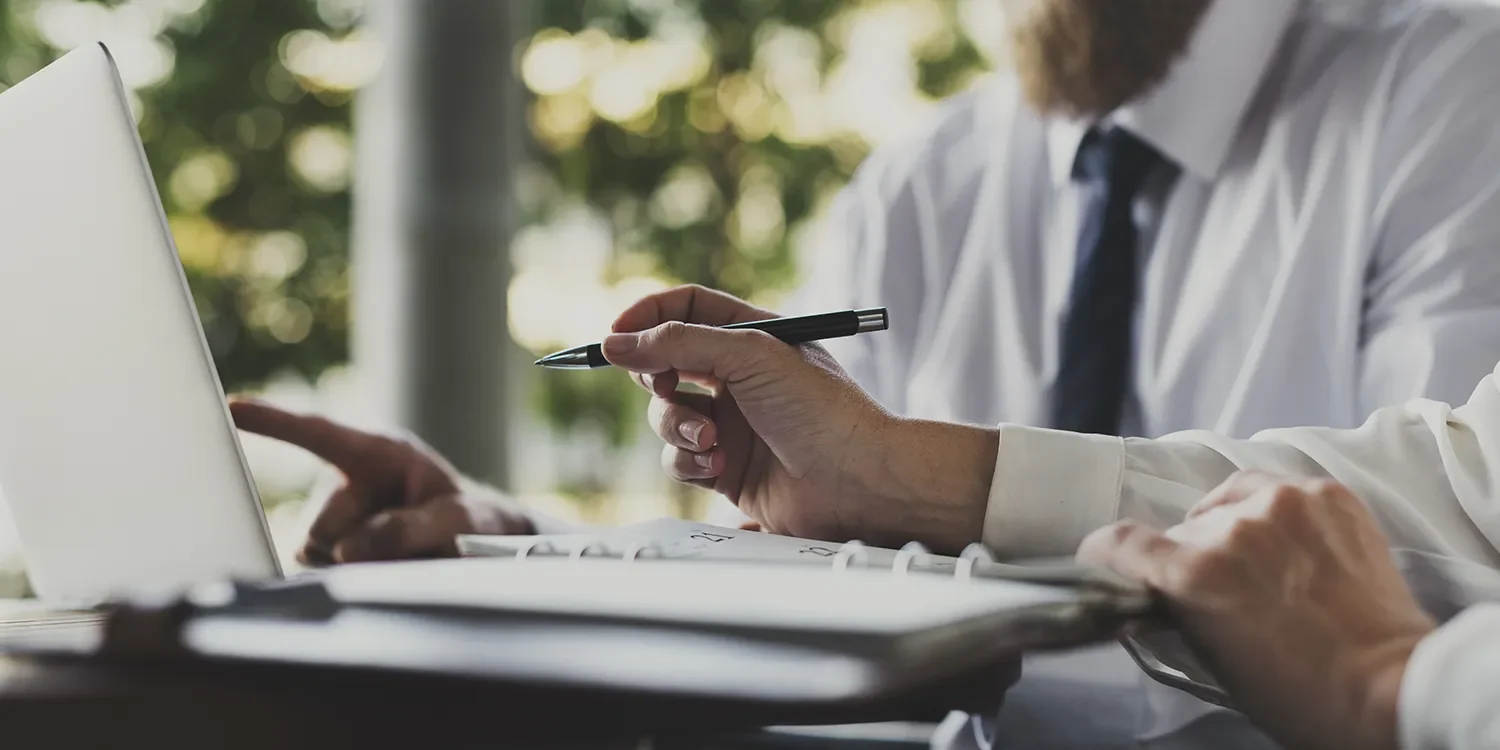 Two people working together at a desk, one is holding a pen and pointing at a laptop screen, with a notebook and papers nearby.