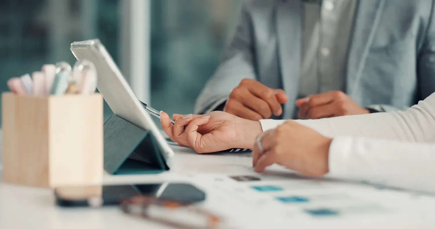 Two people sitting at a desk, looking at a tablet device and discussing, with a pen in one person's hand.
