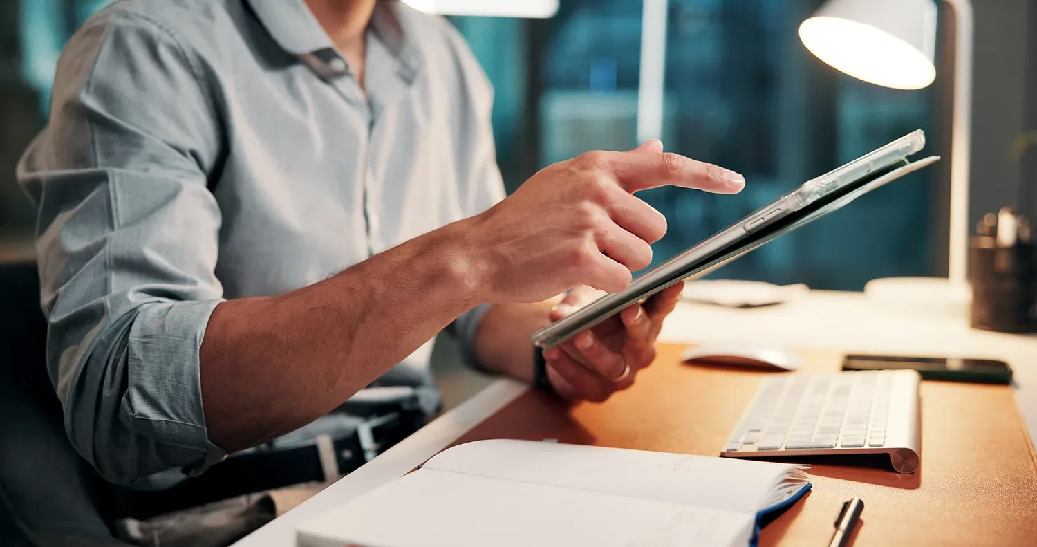 Person in a gray shirt using a tablet at a desk with a keyboard, papers, and a lamp.
