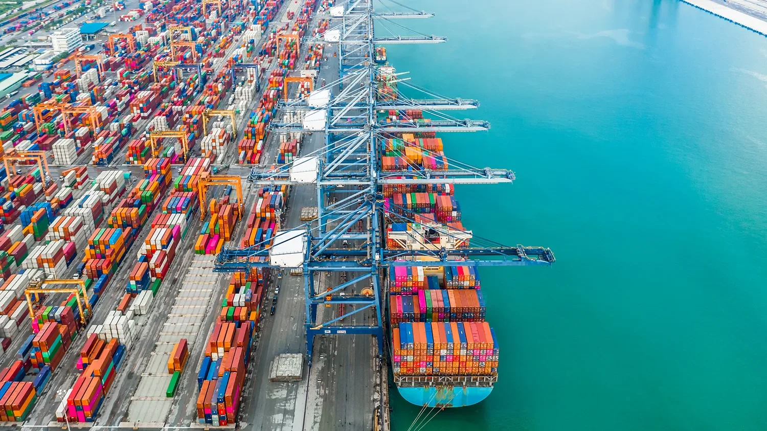 Aerial view of a busy shipping port with numerous colorful containers and large cranes on the dock next to the water.