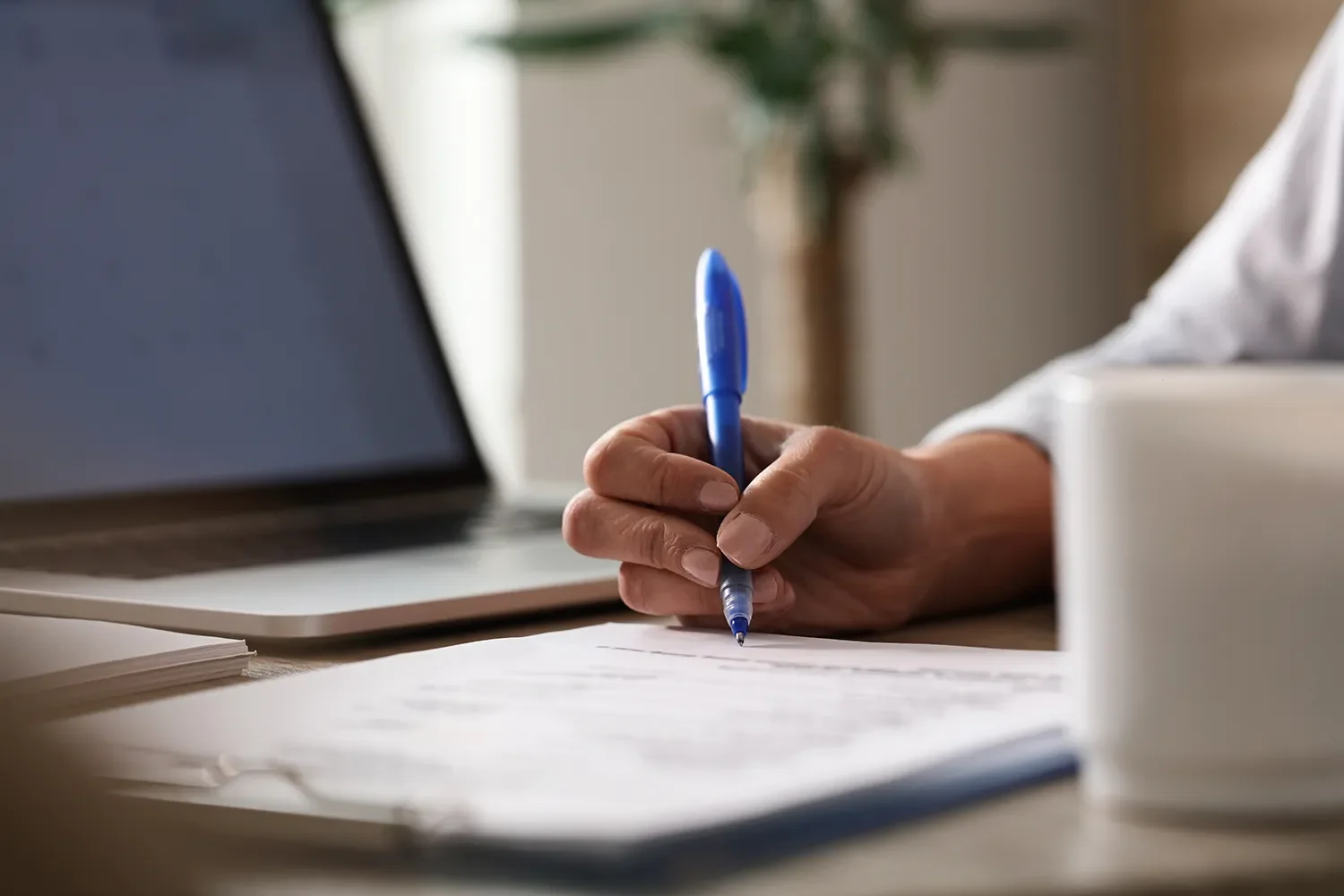 A person's hand holding a blue pen, writing on paper at a desk with a laptop nearby and a stack of white paper or notebook in the foreground.