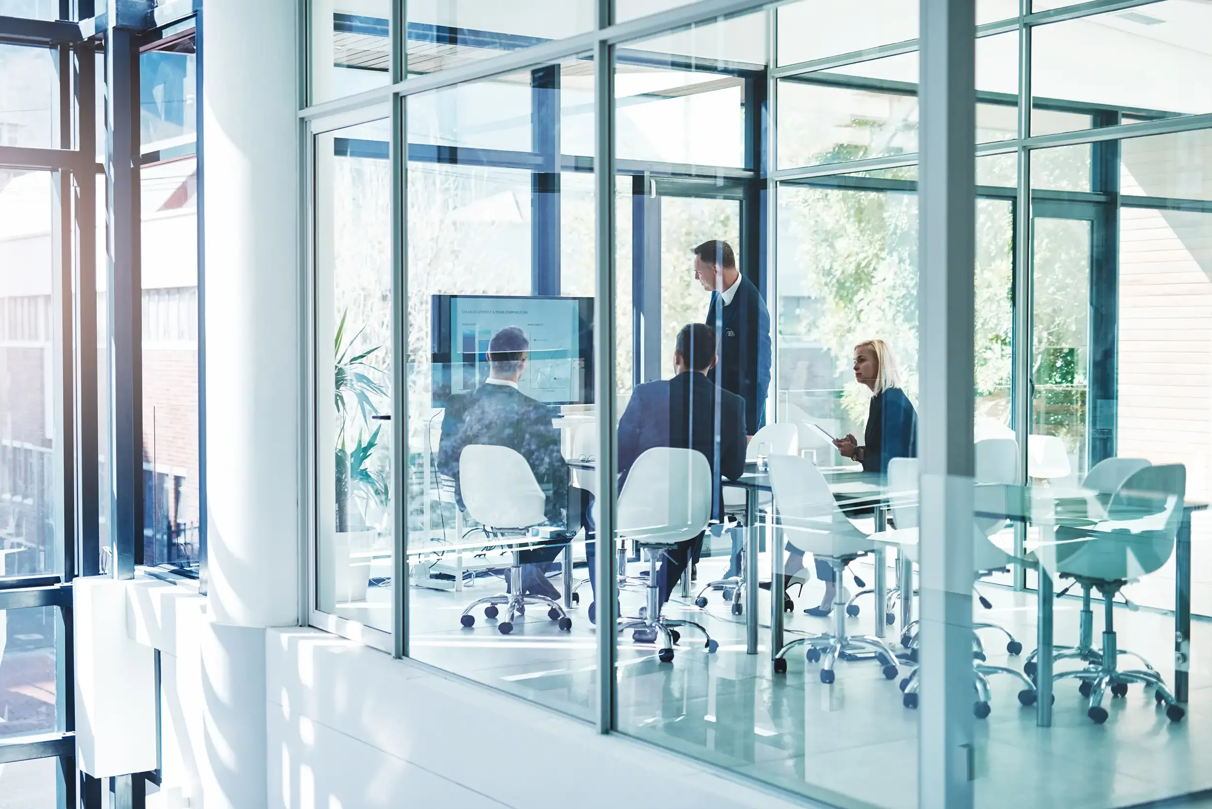 Business meeting in a glass-walled conference room with five people, some standing and some seated, discussing charts on a screen.
