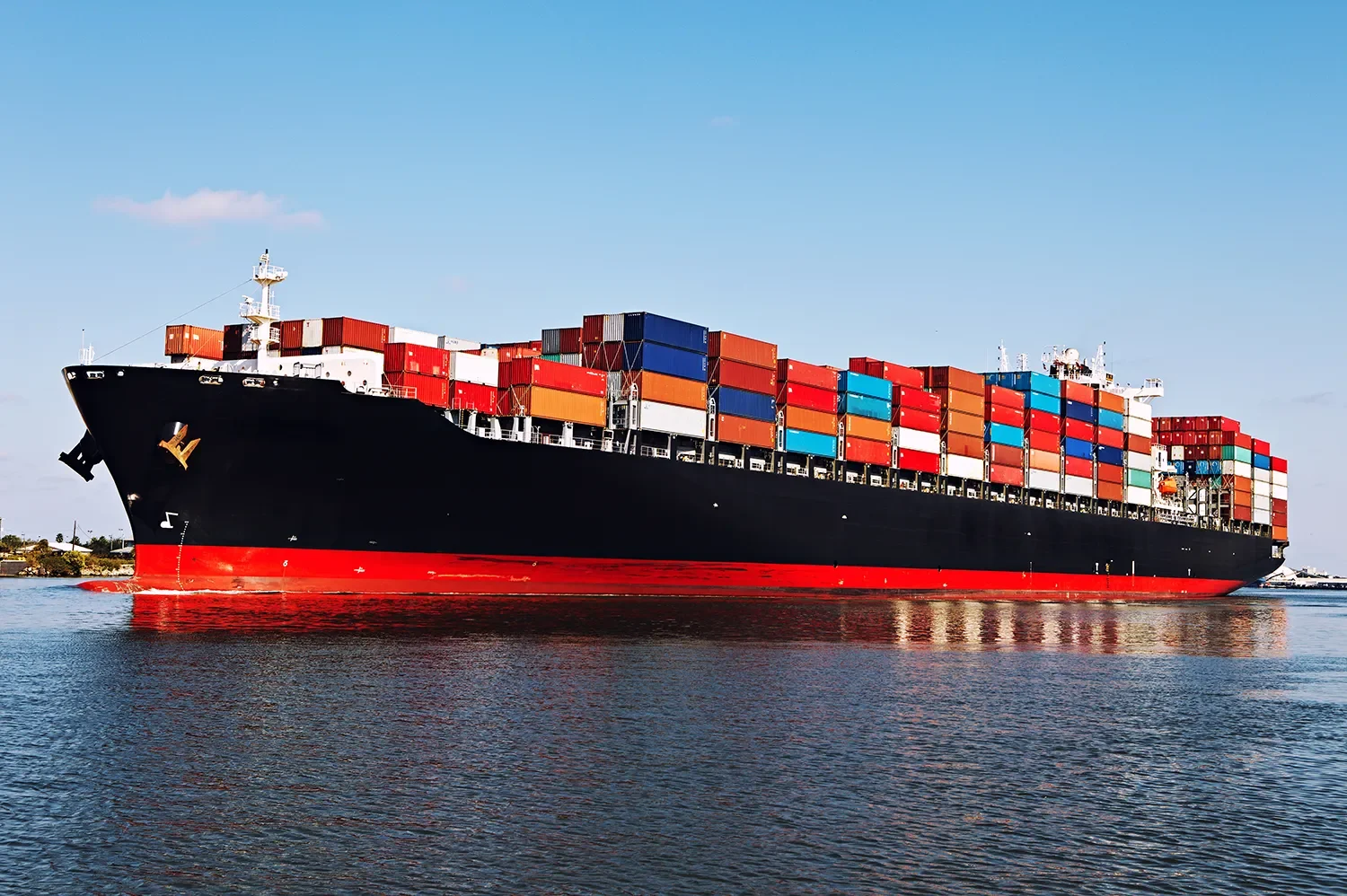 Large cargo ship sailing in calm water, loaded with multicolored shipping containers, under a clear blue sky.