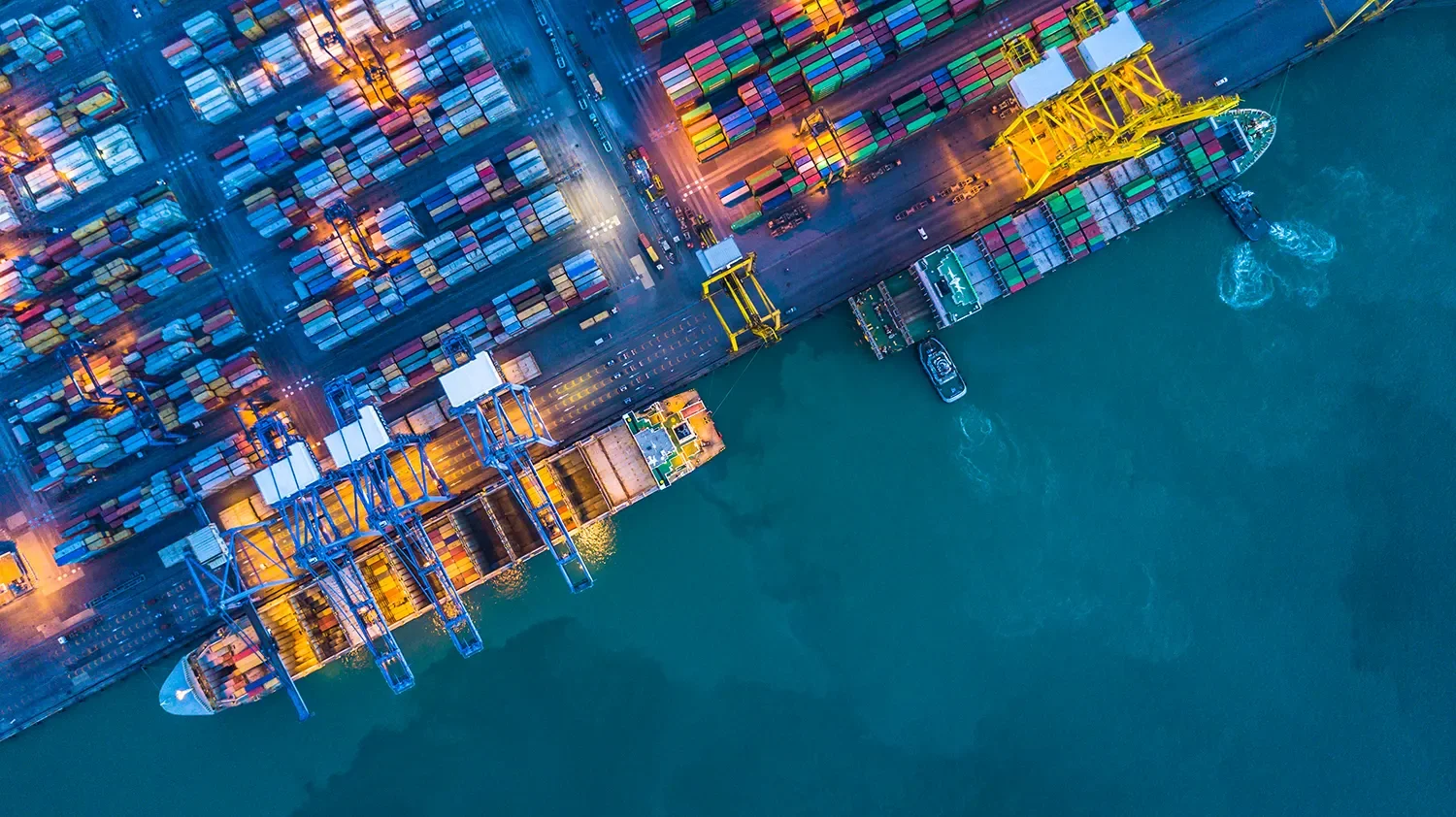 Aerial view of a container port with ships, cranes, and stacked shipping containers, illuminated at dusk.
