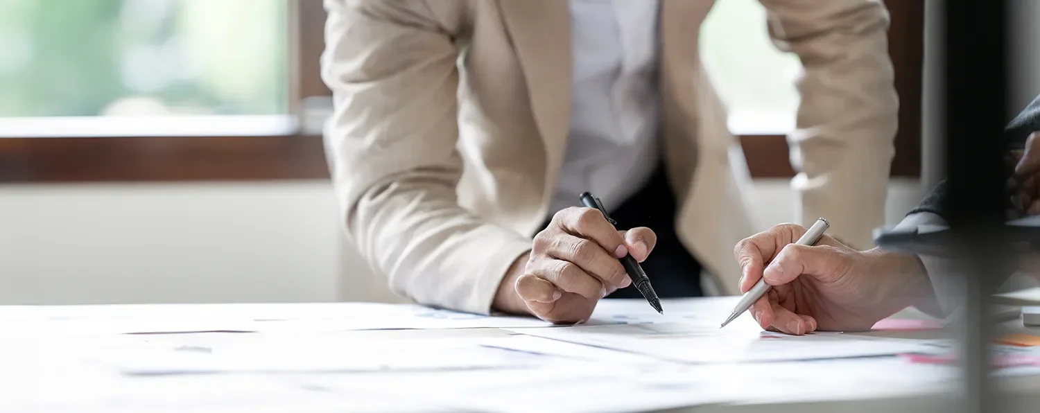 Two people are working on a document or blueprint at a table, with one person pointing with a pen and the other holding a pen, near a window.
