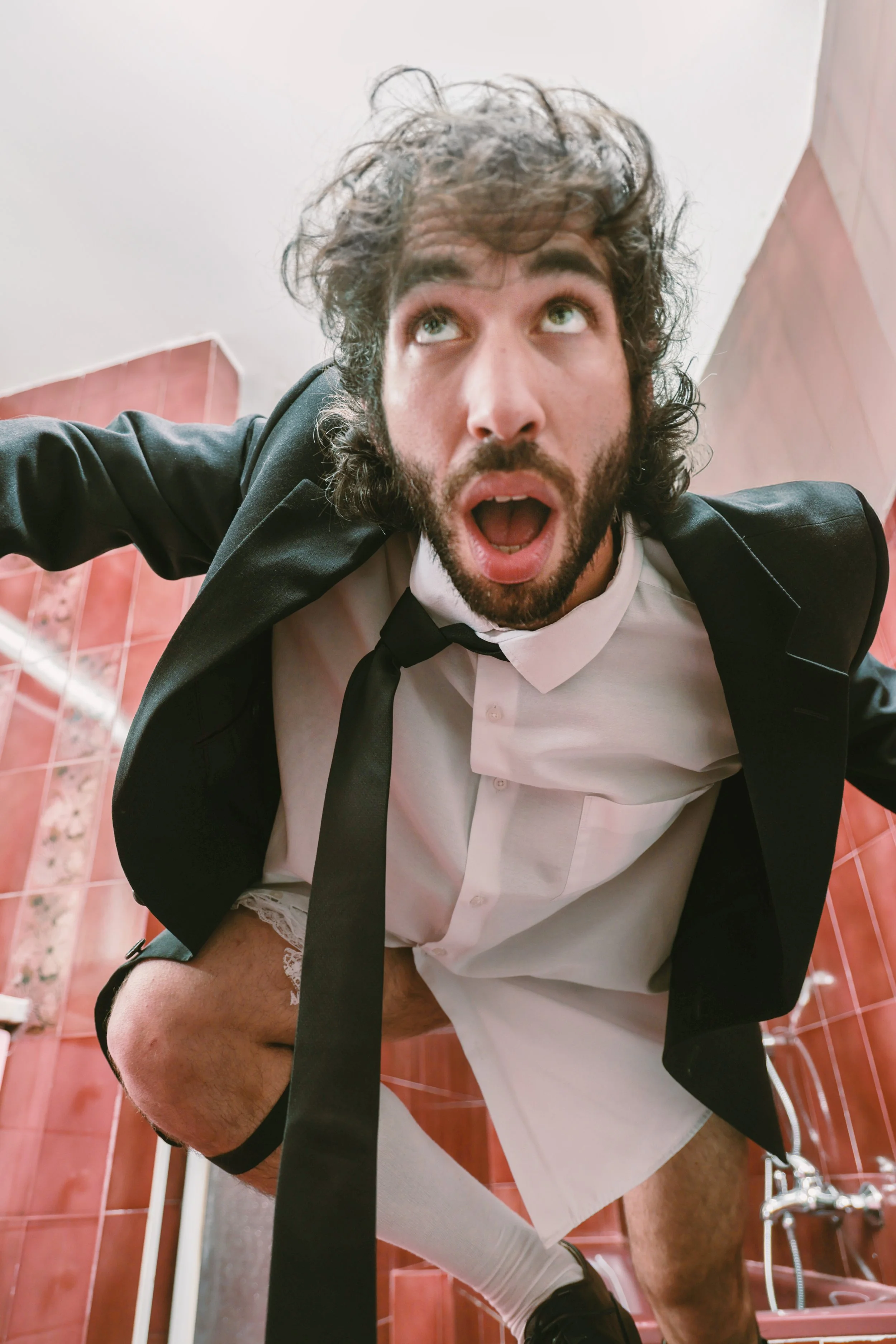 Man with disheveled hair and beard in a bathroom, leaning forward with his mouth open, wearing a white shirt, black blazer, black tie, and white underwear, standing on a toilet with the pants pulled down.