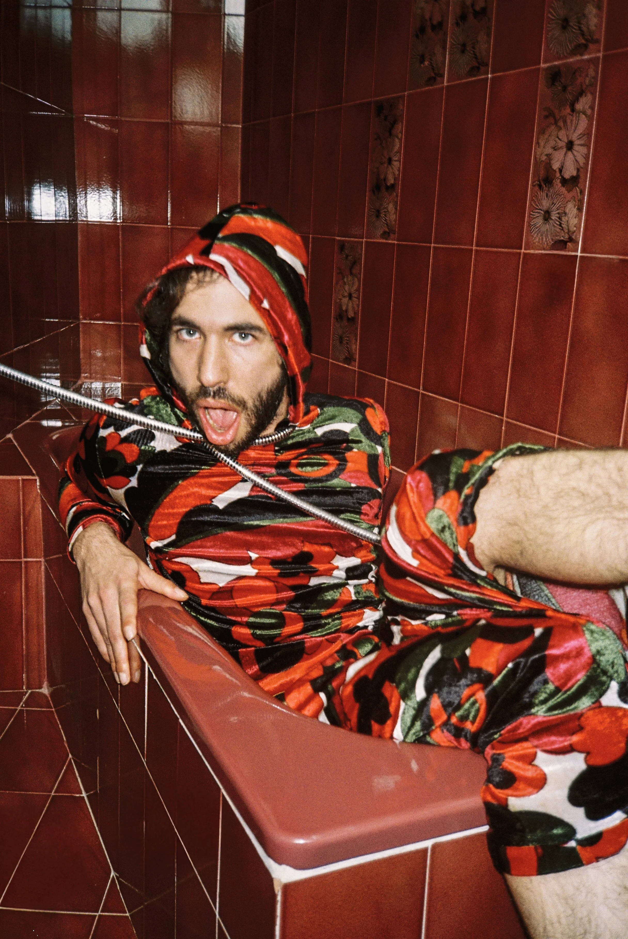 A man in a colorful, floral-patterned outfit, with a matching hood, sitting in a red-tiled bathtub, making a surprised or shocked expression.
