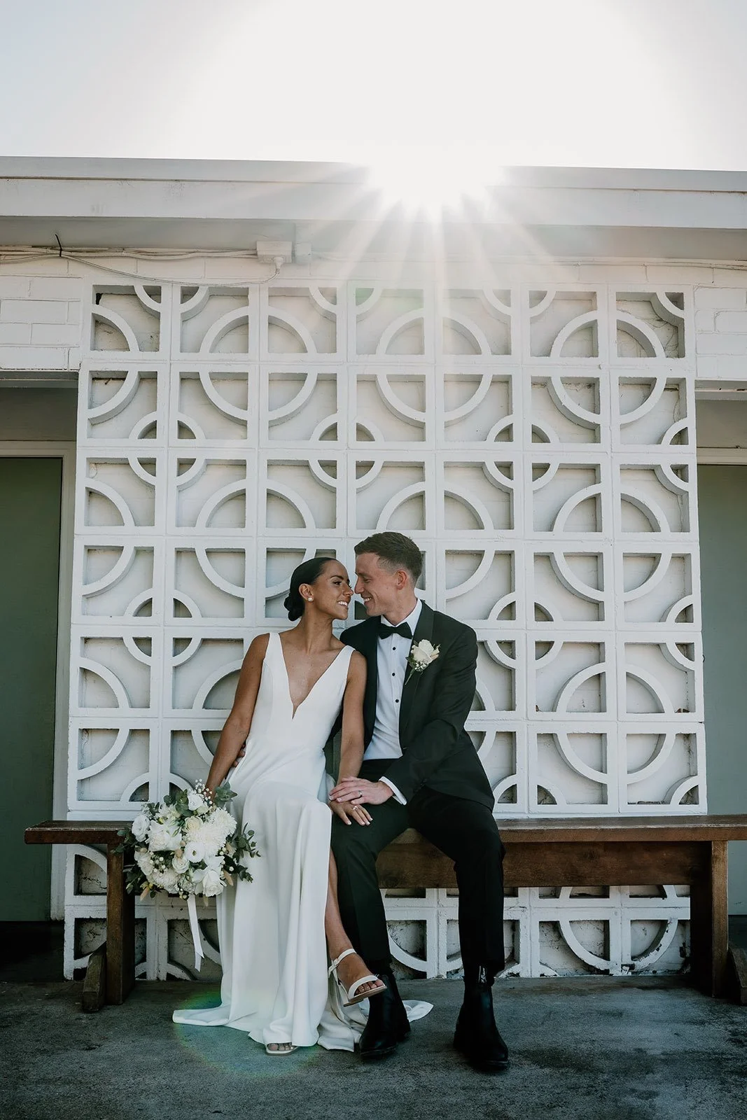 A bride and groom sitting on a wooden bench, smiling and looking at each other, with the bride holding a bouquet of white flowers and the groom wearing a black tuxedo, in front of a decorative white wall with sunlight shining above.