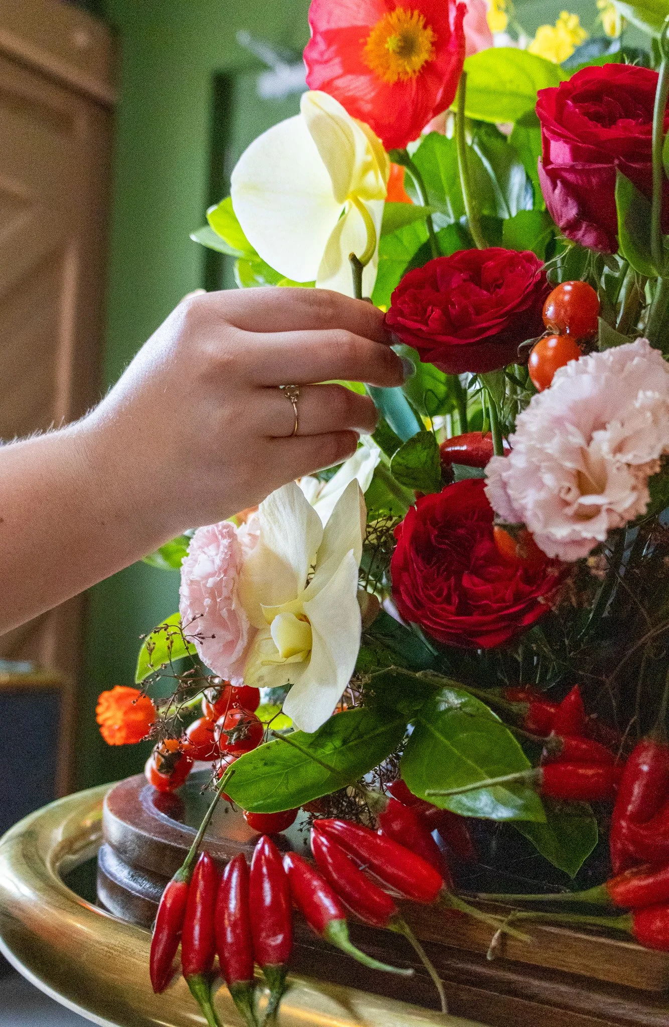 A person's hand arranging a colorful bouquet of flowers, including red and pink roses, white orchids, orange berries, and red chili peppers, on a golden tray.