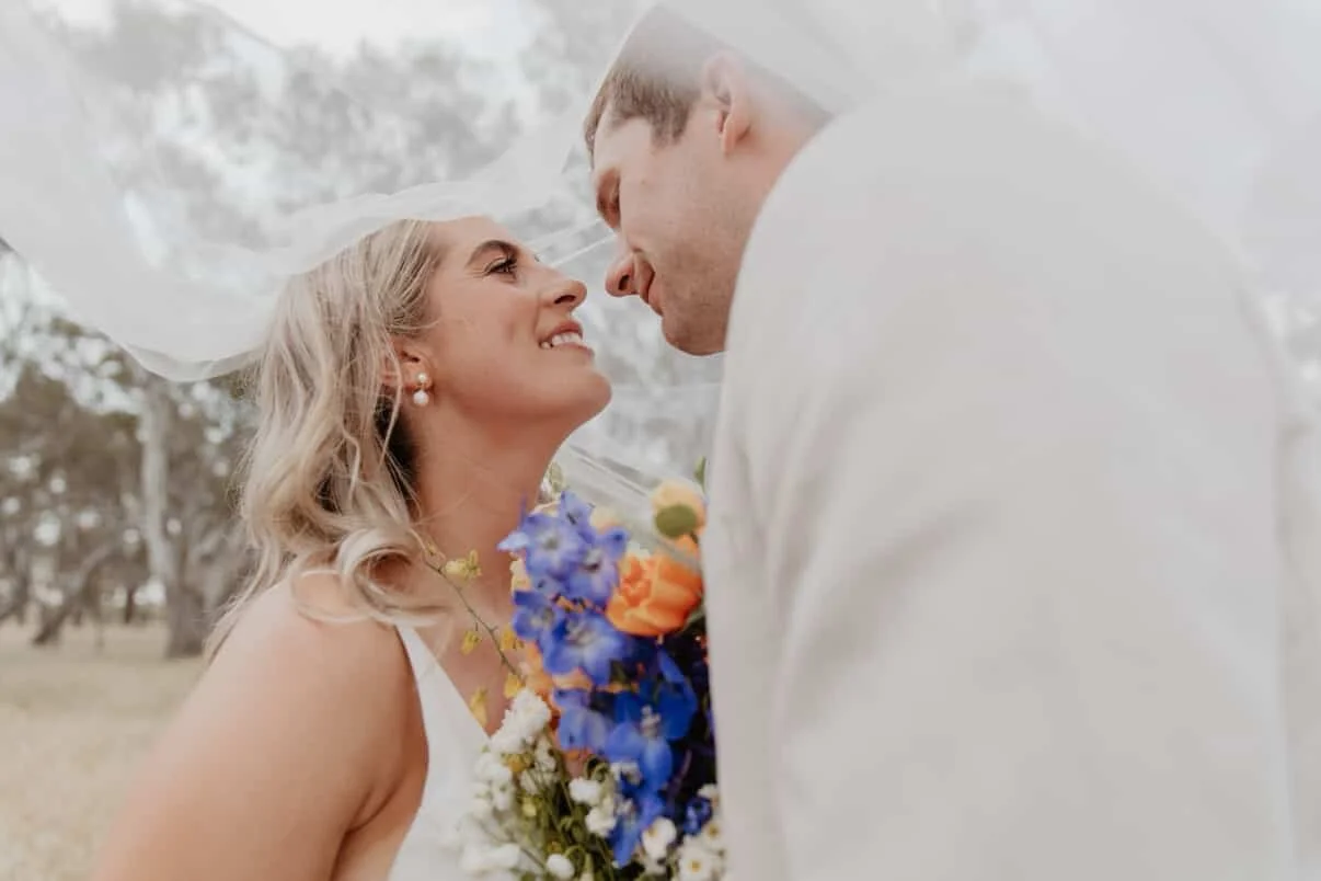 A bride and groom face each other closely outside, holding a bouquet of flowers, with the bride smiling and looking at the groom.