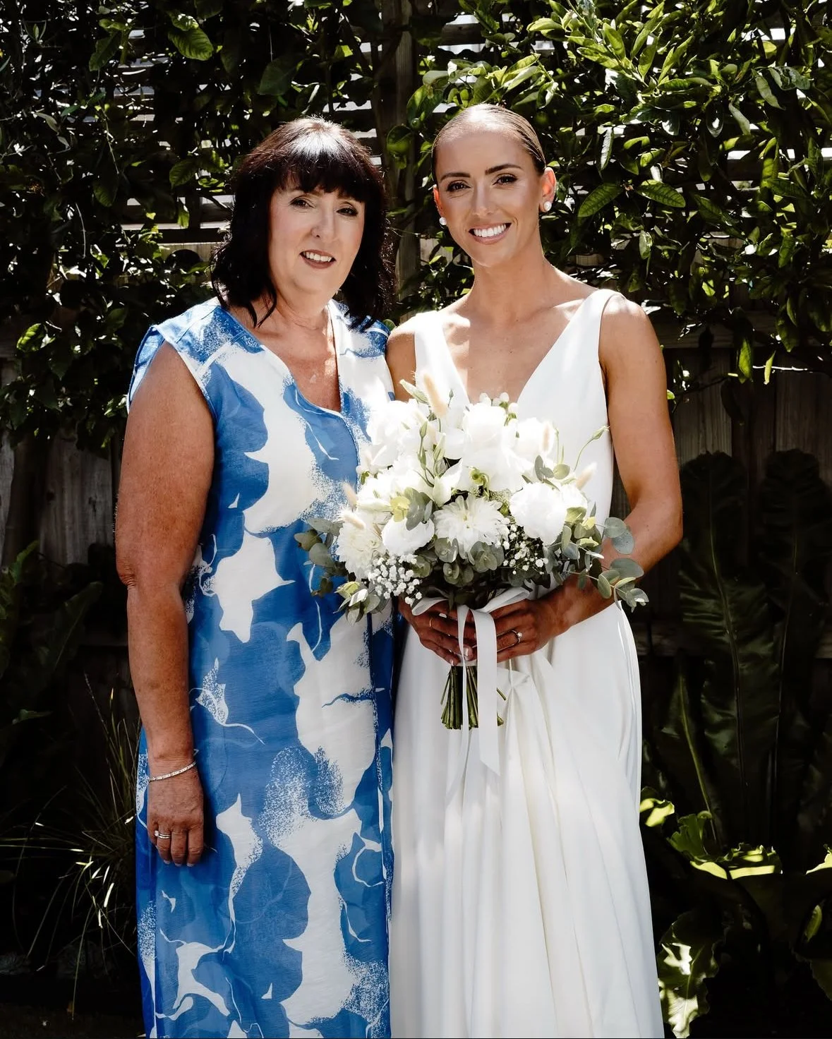 A woman in a white wedding dress holding a bouquet of white flowers, standing next to another woman in a blue and white dress, outdoors with greenery in the background.