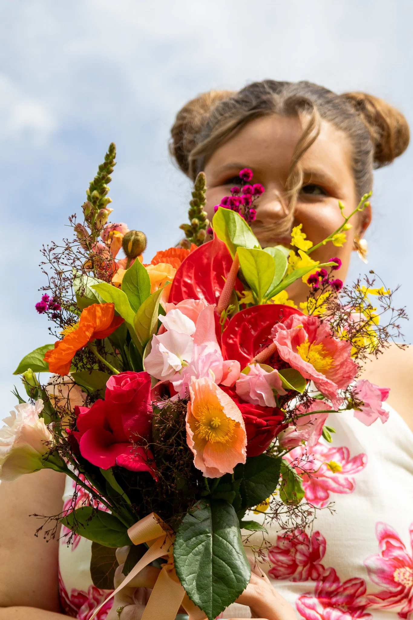 A woman holding a colorful bouquet of flowers, smiling, with a blue sky in the background.