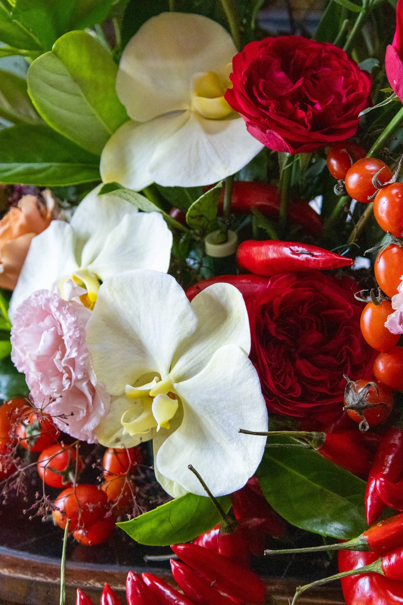 Close-up of a flower arrangement with white orchids, red roses, bright red chili peppers, orange cherry tomatoes, and pink carnations surrounded by green leaves.