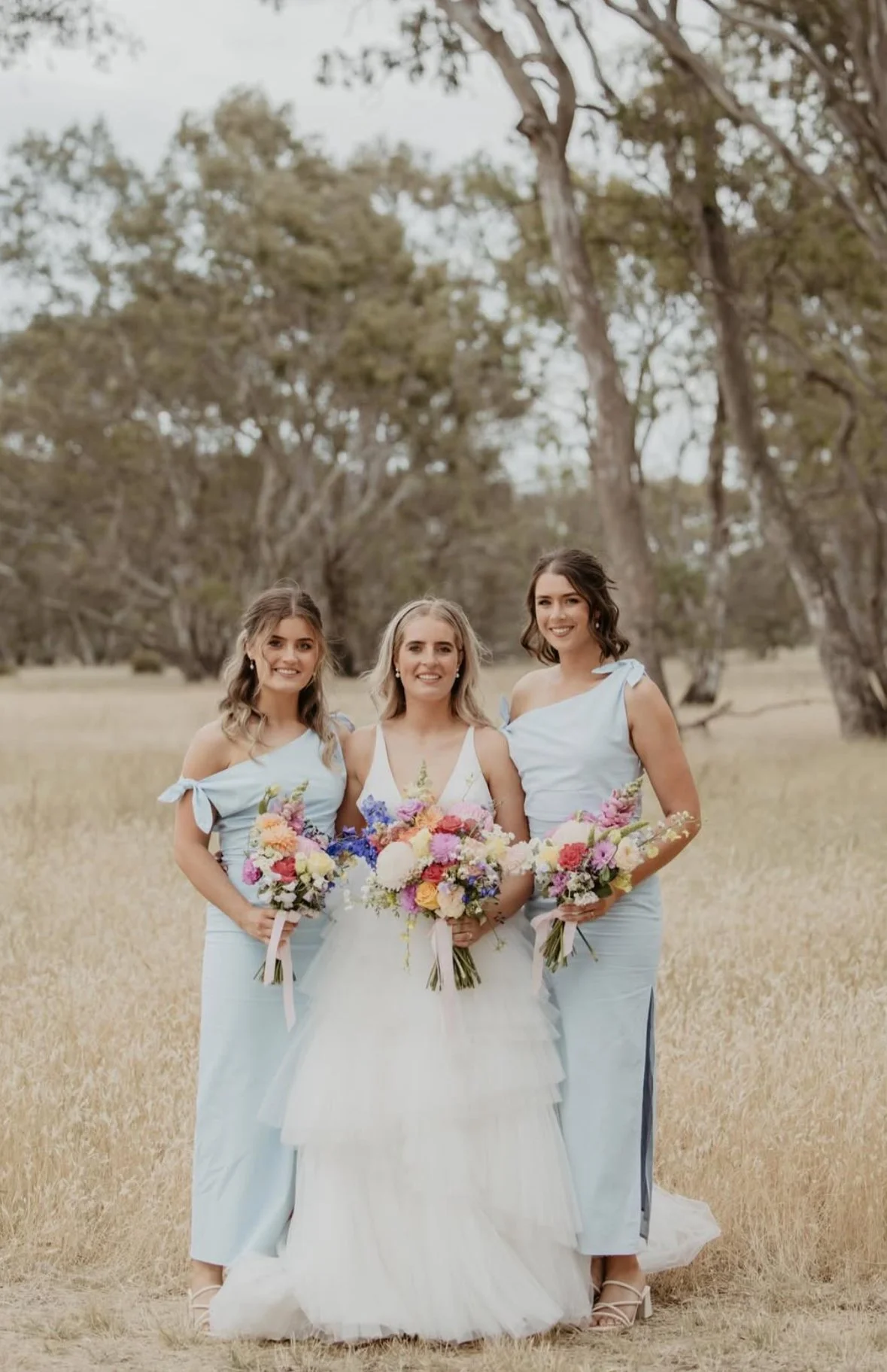 Three women, including a bride in a white wedding dress, standing outdoors in a grassy field with trees in the background, holding bouquets of colorful flowers.