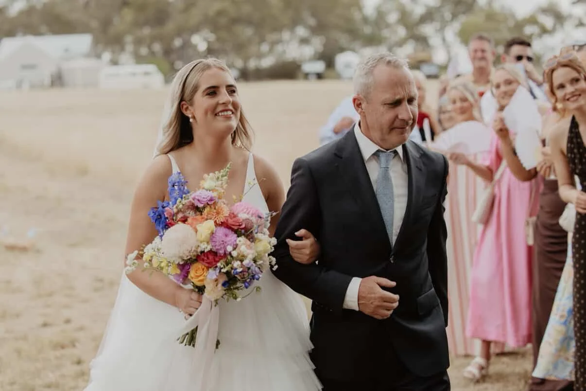 A bride in a white wedding dress holding a bouquet of colorful flowers walking down the outdoor aisle with an older man, surrounded by smiling guests.