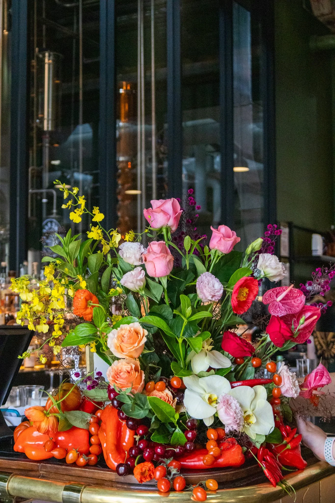 A vibrant bouquet of various flowers, including roses, orchids, and anthuriums, arranged with greenery and red and orange berries, on a floral display table inside a building.
