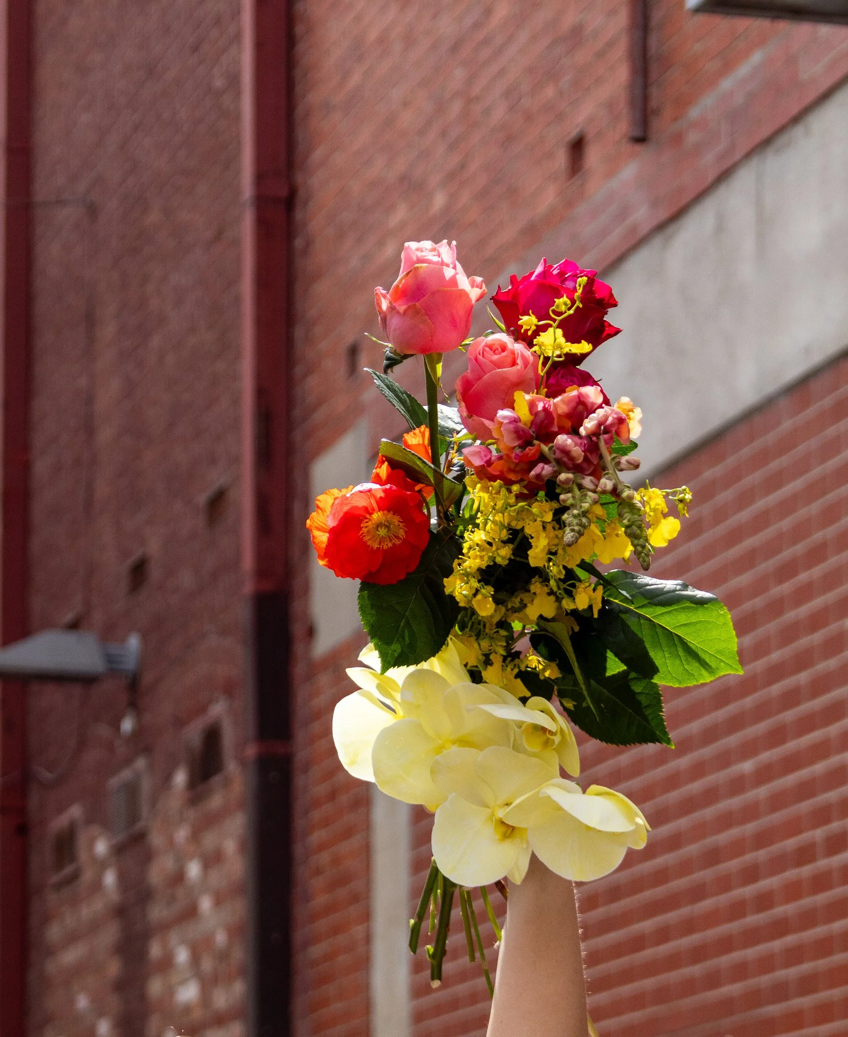 A hand holding a colorful bouquet of flowers against a background of red brick and concrete walls.