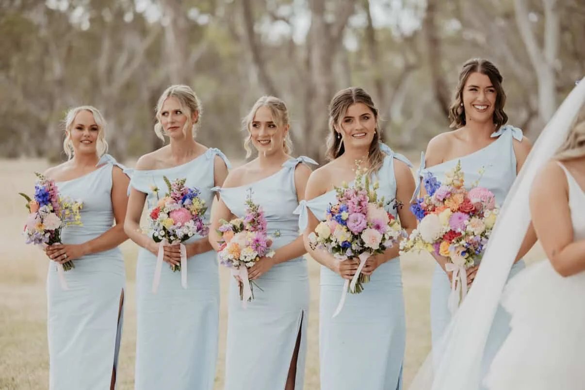Group of bridesmaids in light blue dresses holding colorful bouquets, outdoors in a natural setting.