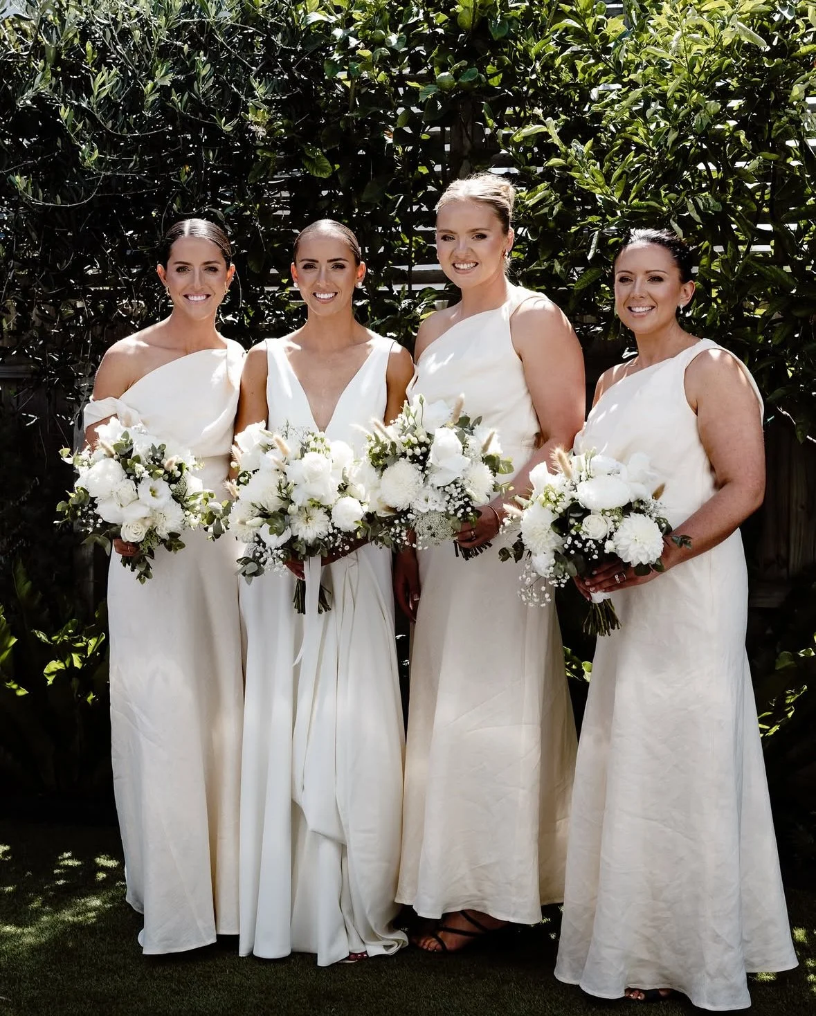 Four women in white dresses holding bouquets of white flowers, standing outdoors with leafy background.
