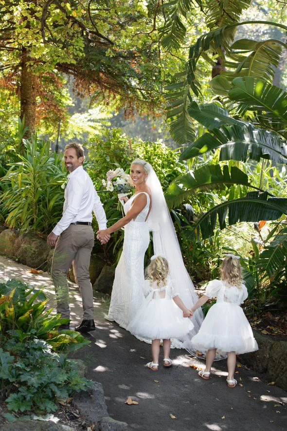 A newlywed couple holding hands and smiling, walking in a lush green garden, with two young girls in white dresses holding hands and walking ahead of them.