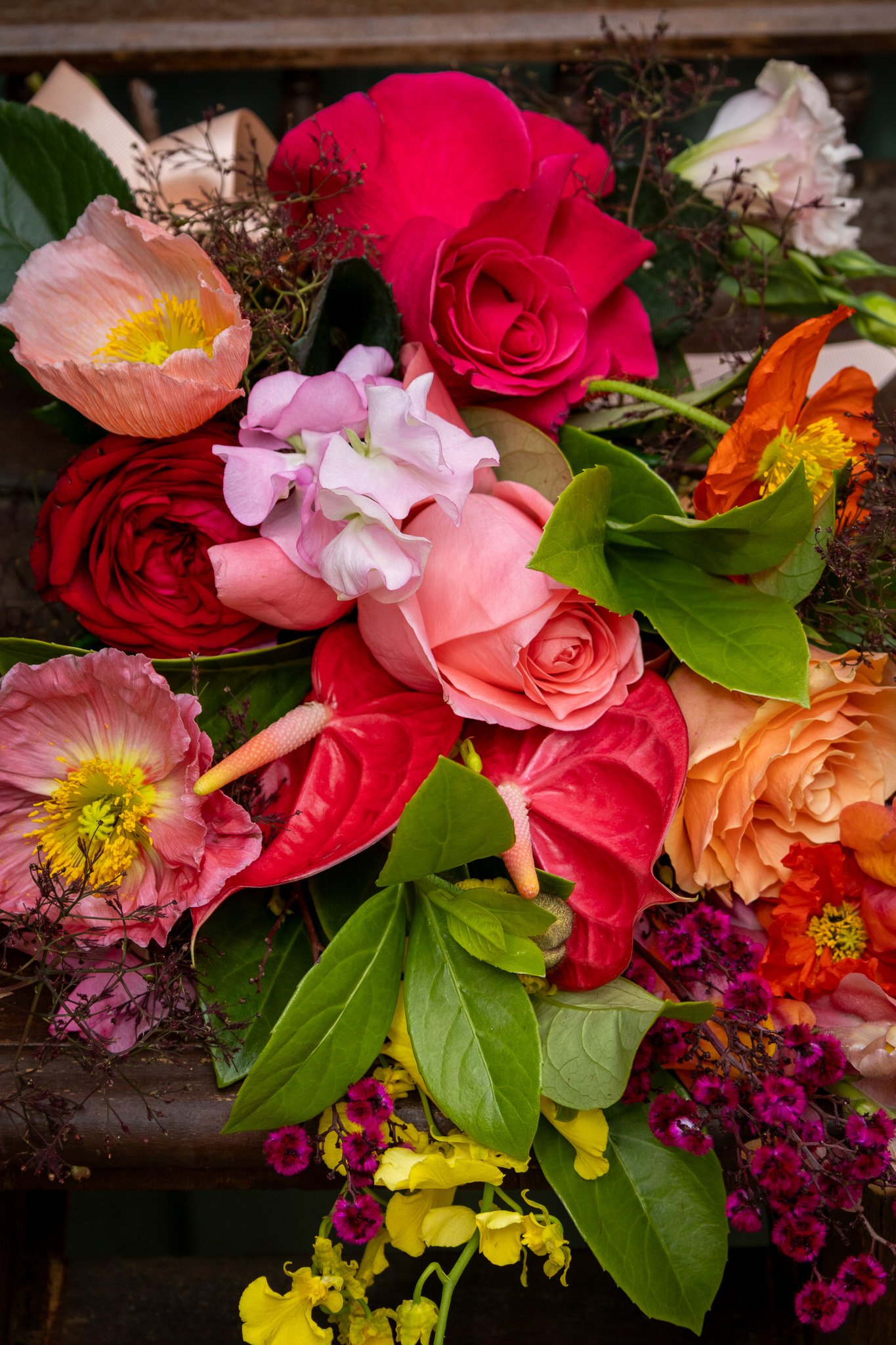 Colorful bouquet of various flowers including roses, anthuriums, anemones, and sweet peas with green leaves, arranged on a rustic wooden surface.