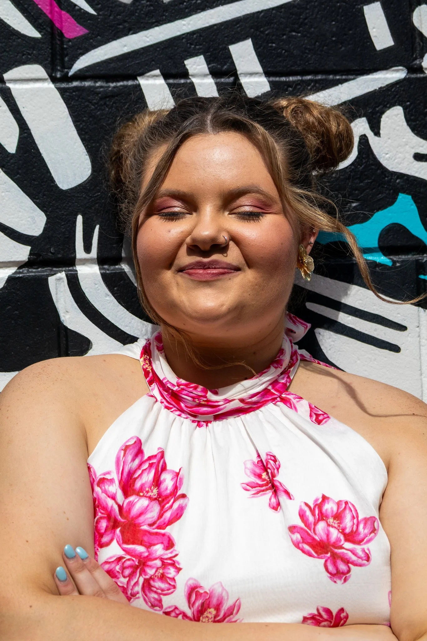 A woman with her eyes closed, smiling, with her arms crossed, in front of a black and white mural with some pink and blue accents, wearing a white dress with pink floral patterns and matching pink scarf-like collar, and golden earrings.