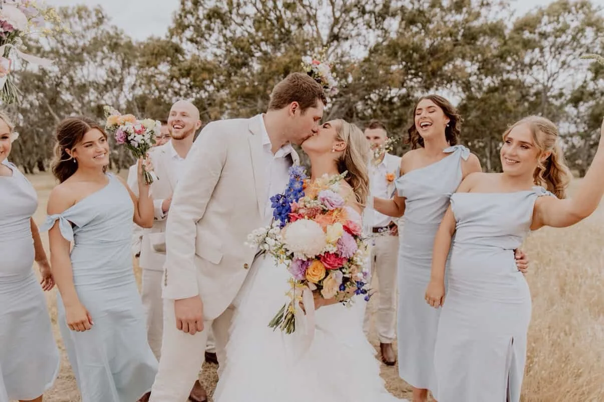 A bride and groom kiss outdoors with a wedding party of six women and two men surrounding them, all dressed in light-colored attire, holding flowers, in a field with trees in the background.
