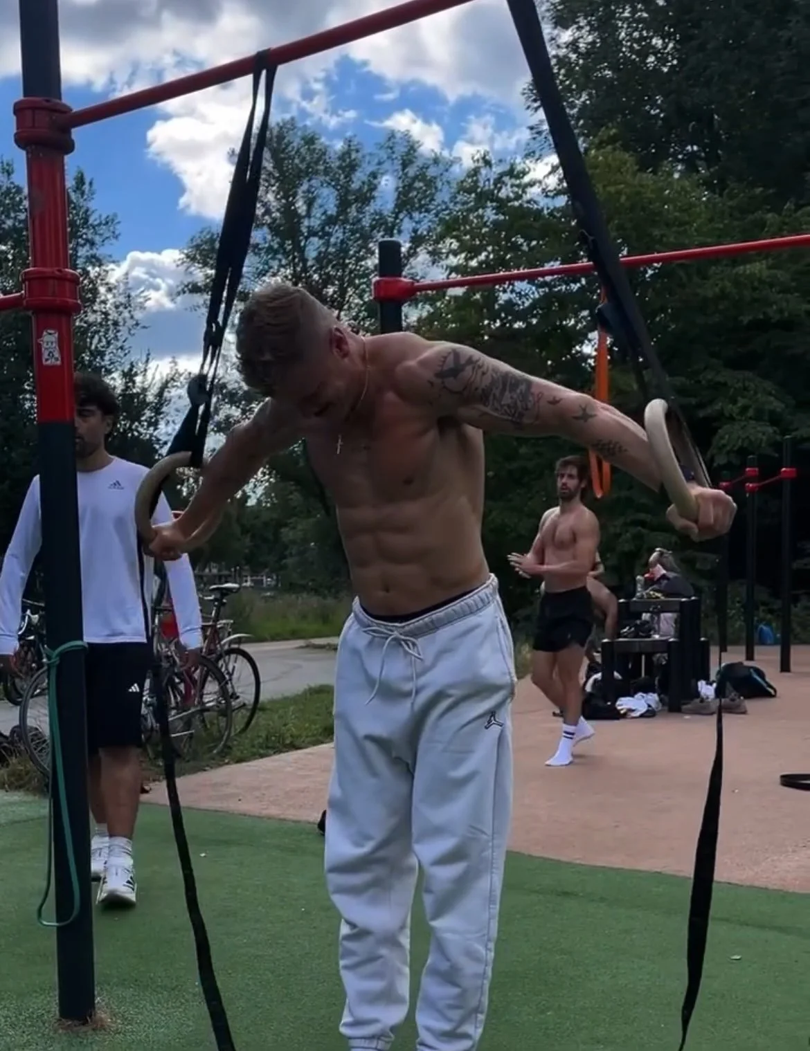 A shirtless man with tattoos on his arms is using gymnastic rings on an outdoor fitness structure, with a park and blue sky with clouds in the background.