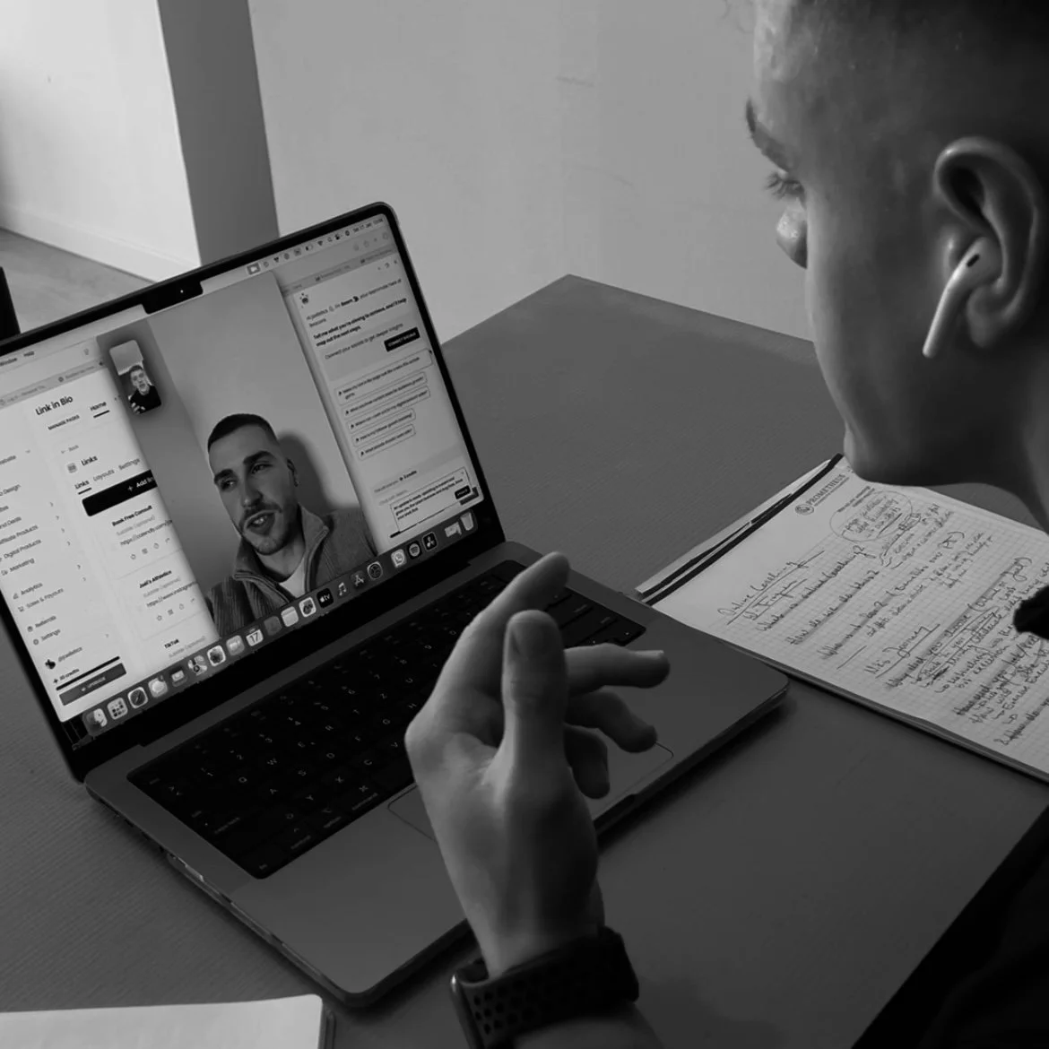 Person having a video call on a laptop with another man, sitting at a desk with handwritten notes.