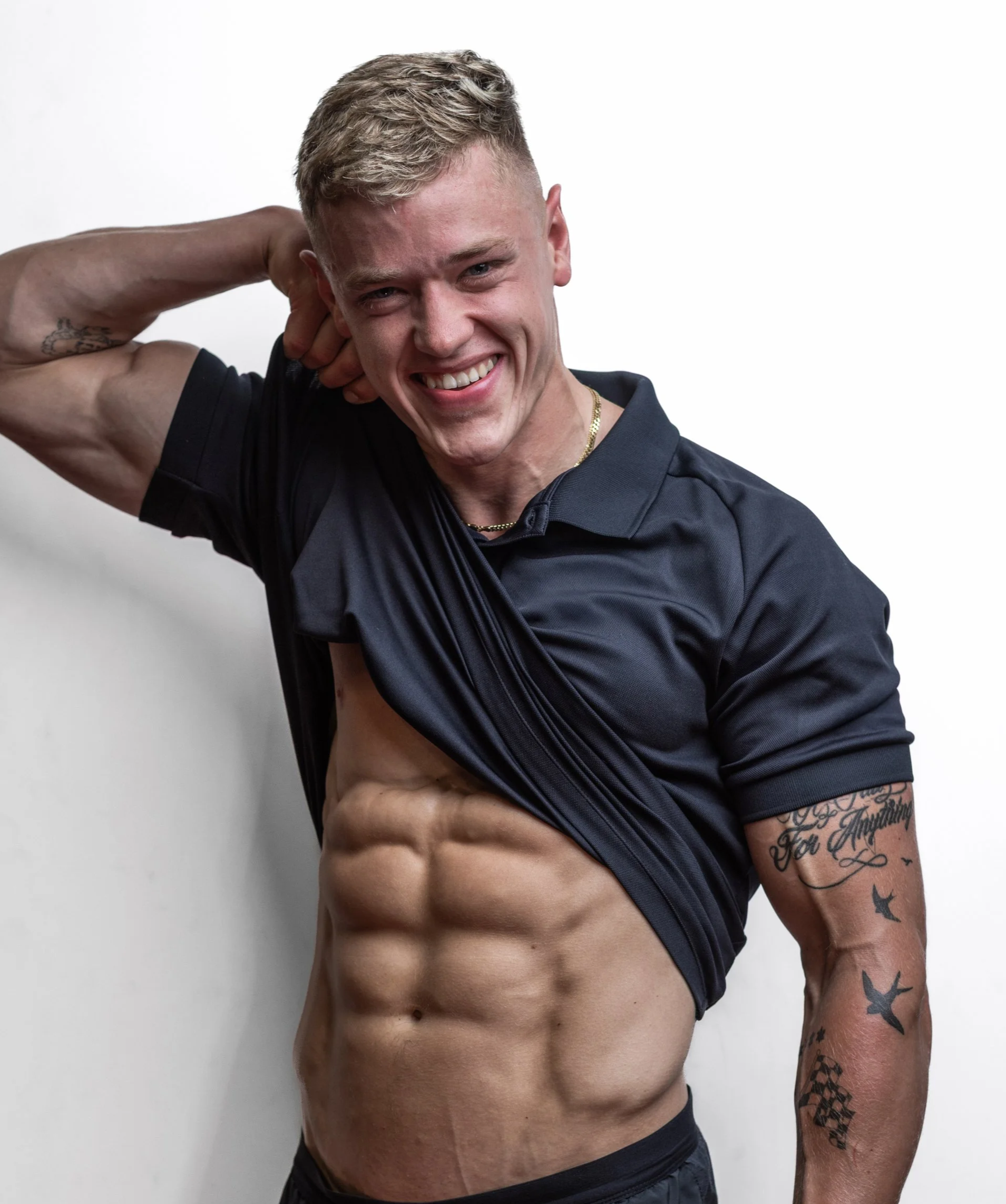 A young man with a muscular, defined abdomen is lifting his shirt to show his abs, while smiling and posing against a plain white background.