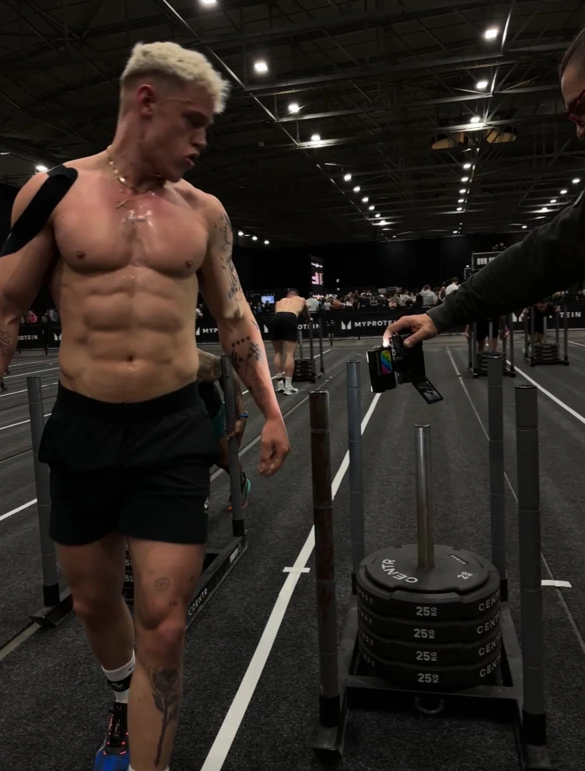 A shirtless male athlete with tattoos on his arms and leg, with bleach blonde hair, at a bodybuilding or fitness event, standing on a running track in an indoor arena, with a trainer or official holding a phone nearby.