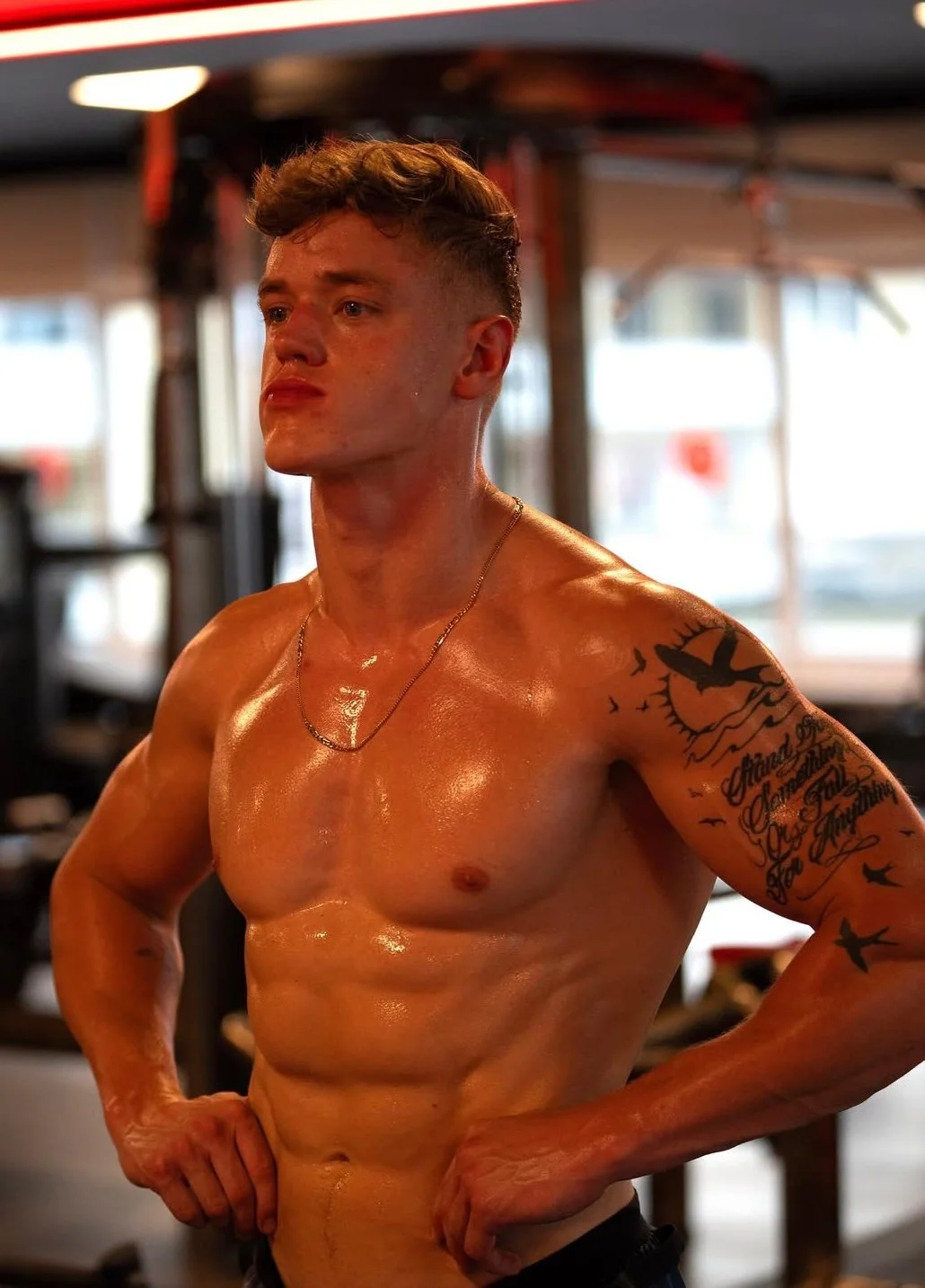 A shirtless muscular young man with short hair and a tattoo on his left arm, standing in a gym with a focused expression.
