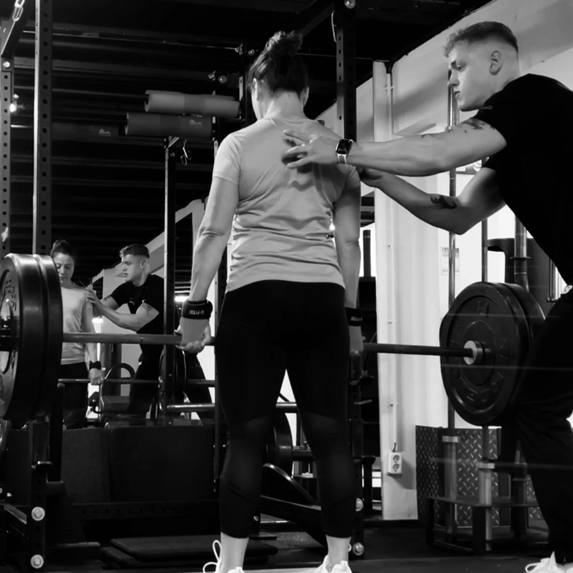 A woman lifting a barbell during a weightlifting workout, with a trainer assisting her in a gym.
