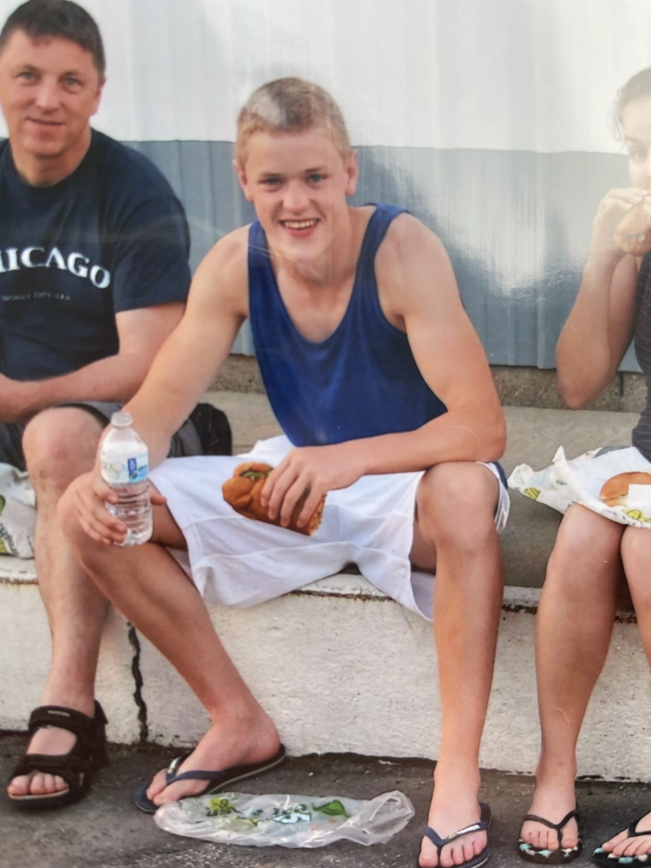 Young man sitting on a curb, holding a water bottle and a hot dog, smiling at the camera, wearing a blue tank top and white shorts. Two other people are partially visible on either side.