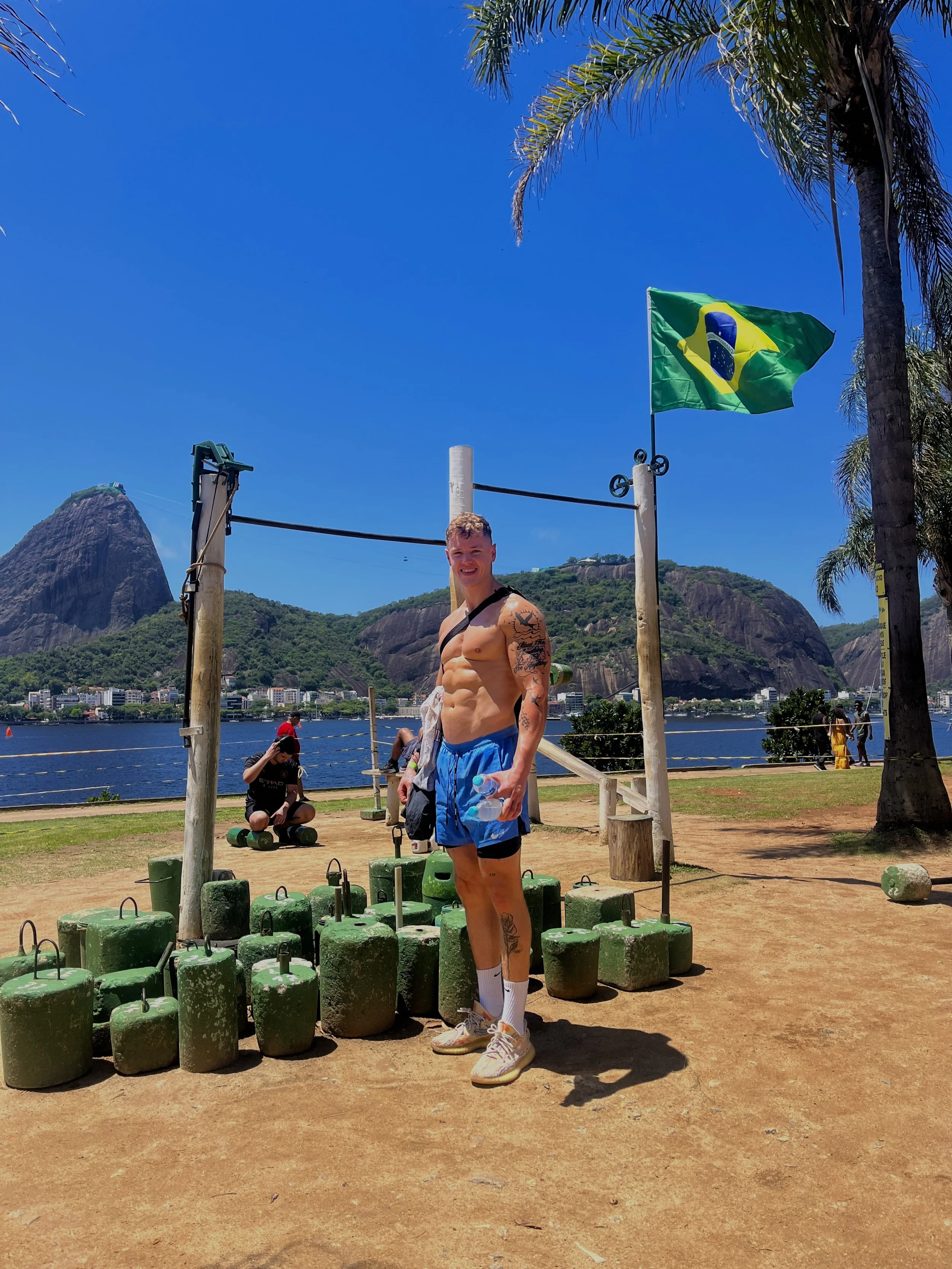 A muscular shirtless man smiling at the camera, standing outdoors near a body of water, with mountains in the background, a Brazilian flag above him, and green weights on the ground.