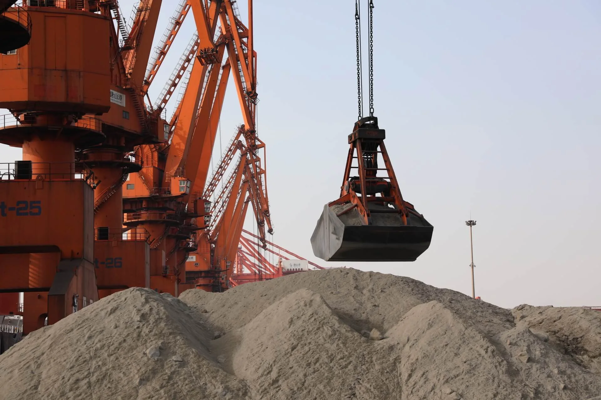 A construction site with a large crane lifting a black and gray container over piles of sand or gravel, with a clear sky in the background.
